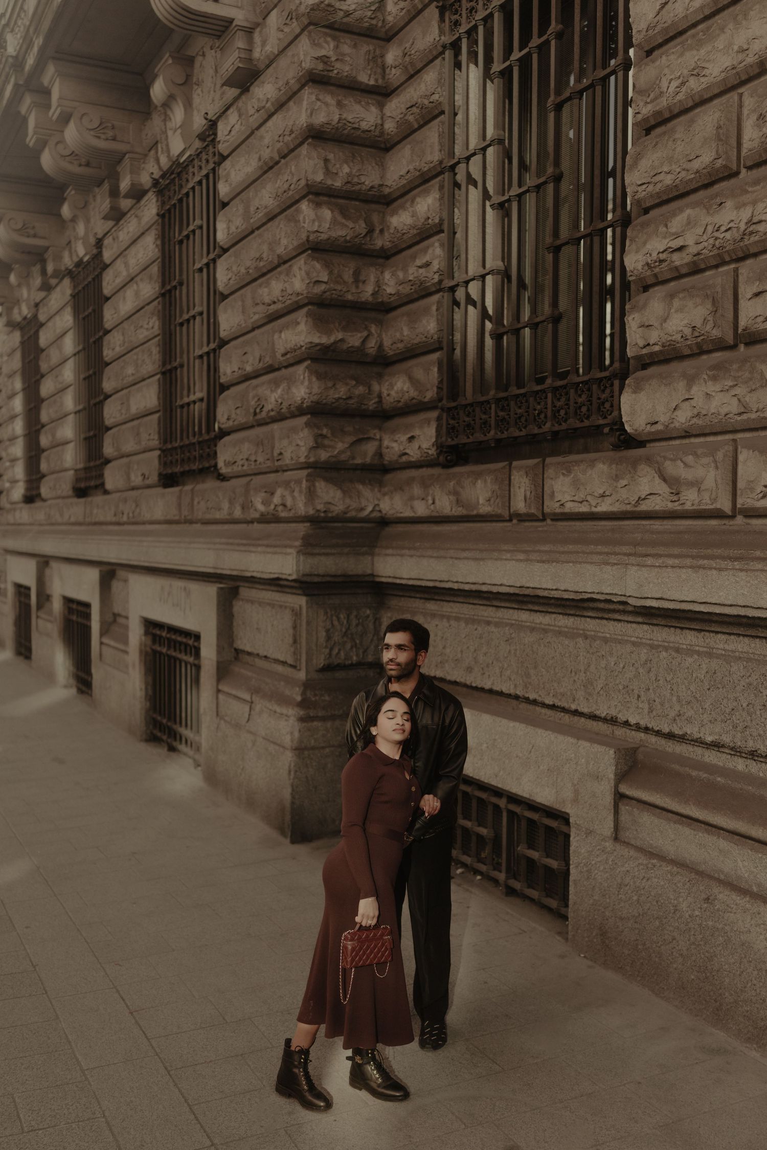 Stylish couple embracing in front of a classic stone building with barred windows.