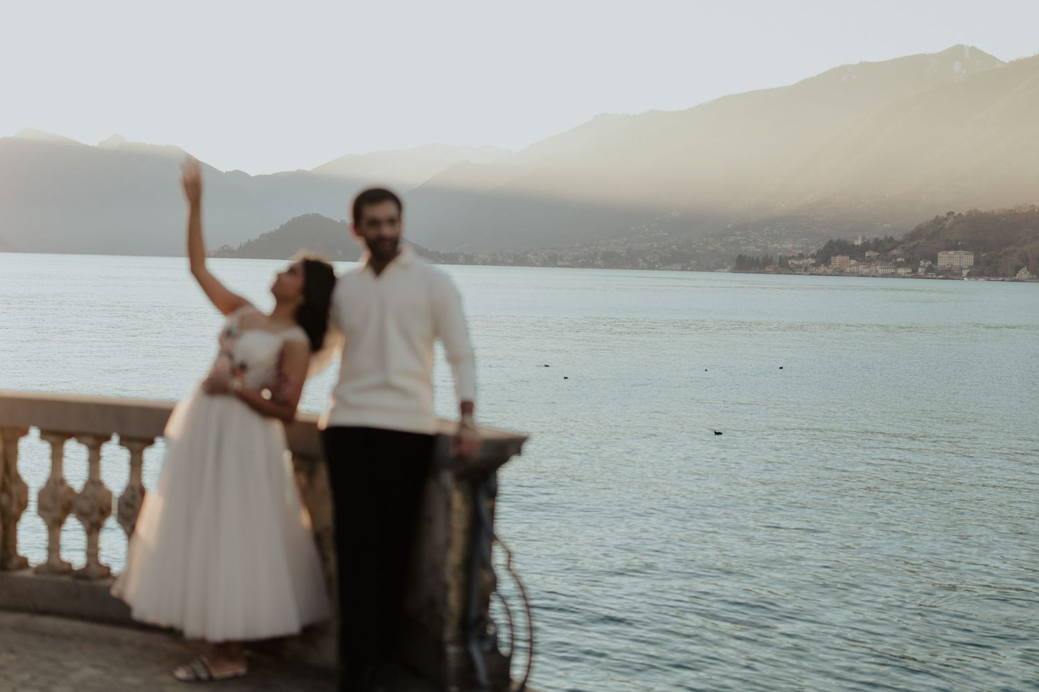 A couple in wedding attire stands by a lakeside balustrade with mountains in the background.