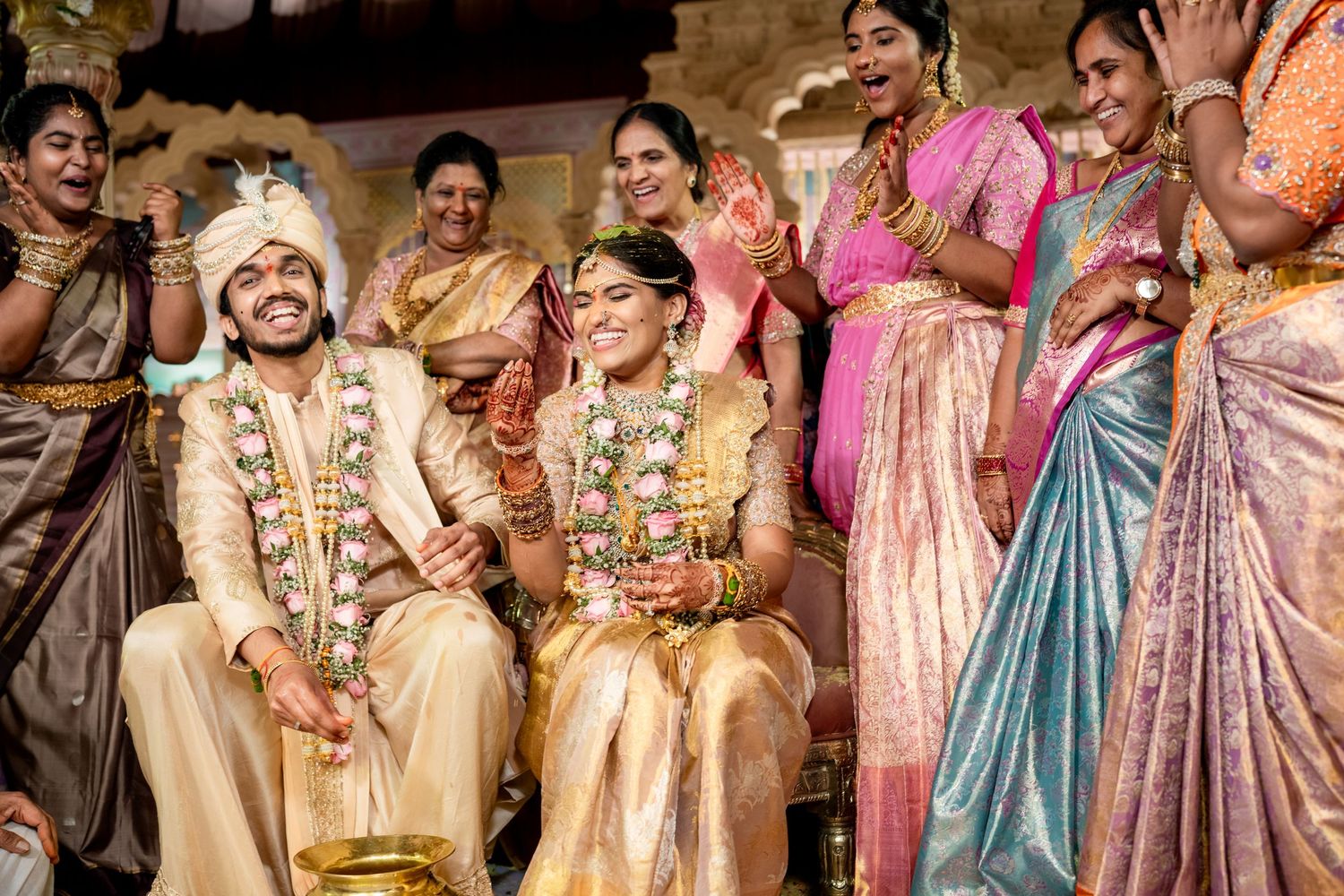 Indian bride and groom exchange garlands joyfully surrounded by family at traditional wedding ceremony.