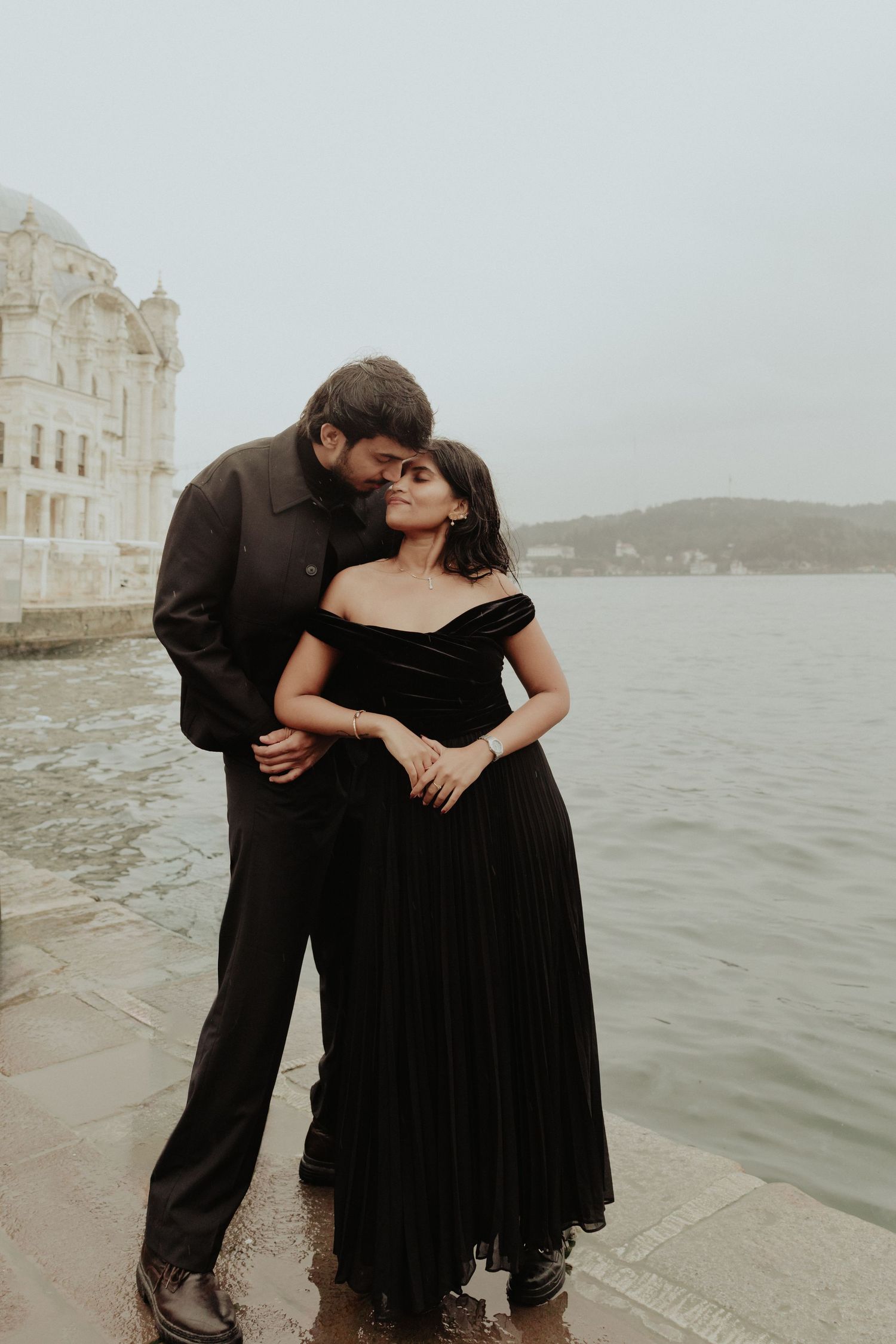 A romantic couple in black attire embraces by the Bosphorus waterfront near Ortakoy Mosque in Istanbul.