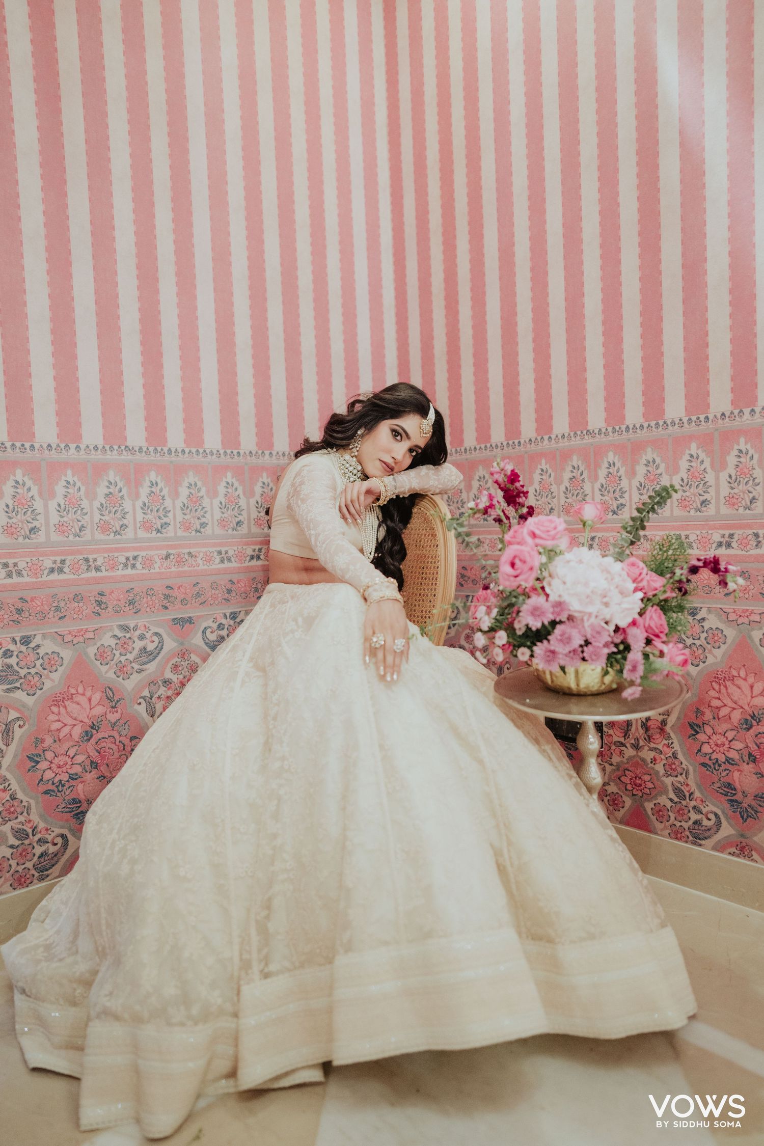 Bride in ivory embroidered ball gown lehenga poses elegantly beside pink floral arrangement against striped wall.