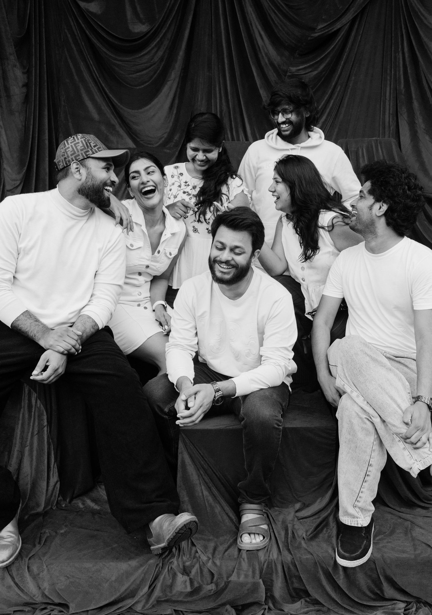 Black and white photo of a joyful group of eight friends laughing together against a draped fabric backdrop.