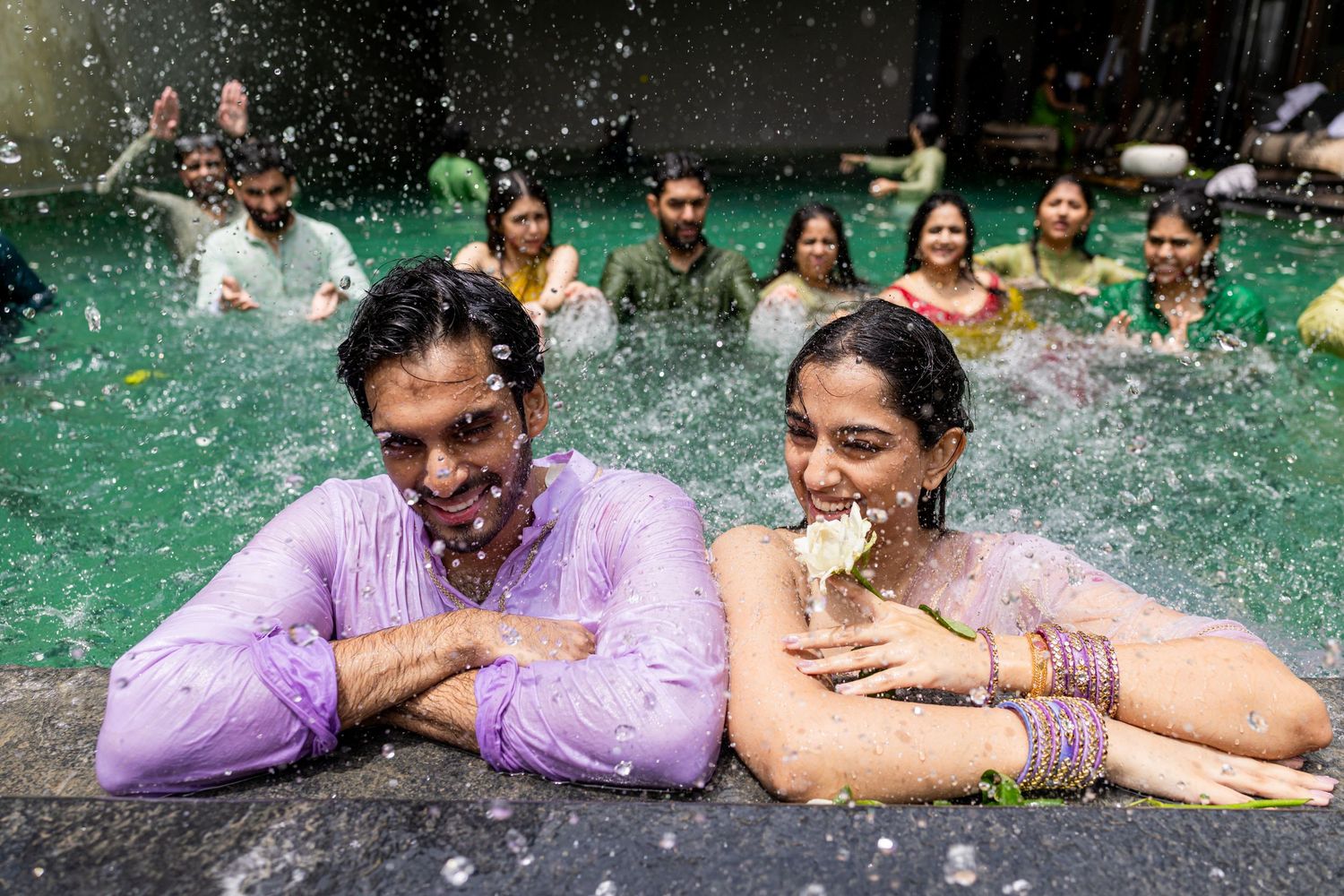 Couple smiling in a green pool surrounded by guests during a vibrant Indian celebration.