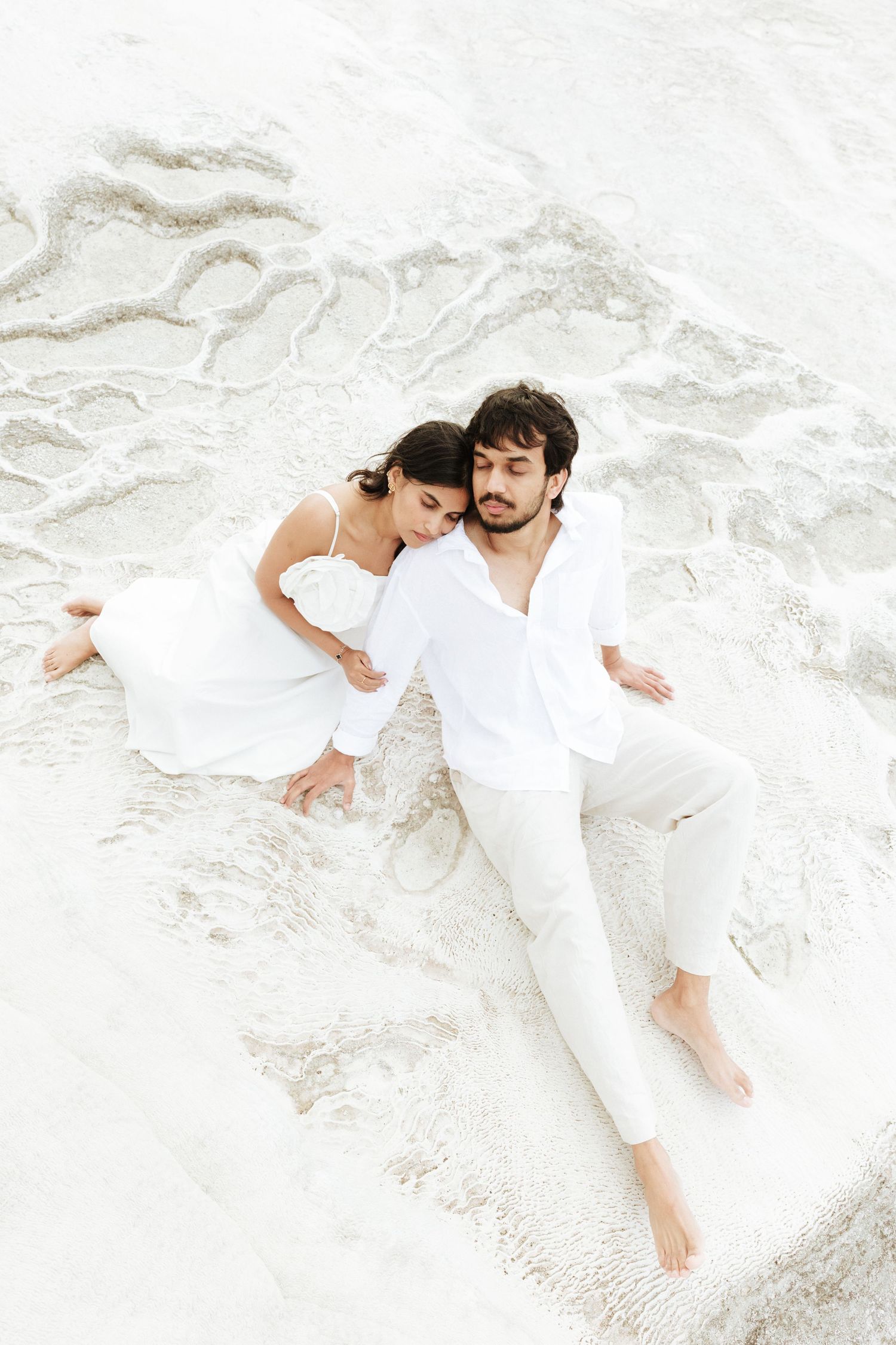 Couple in white outfits lying together on white sand beach, sharing a romantic peaceful moment.
