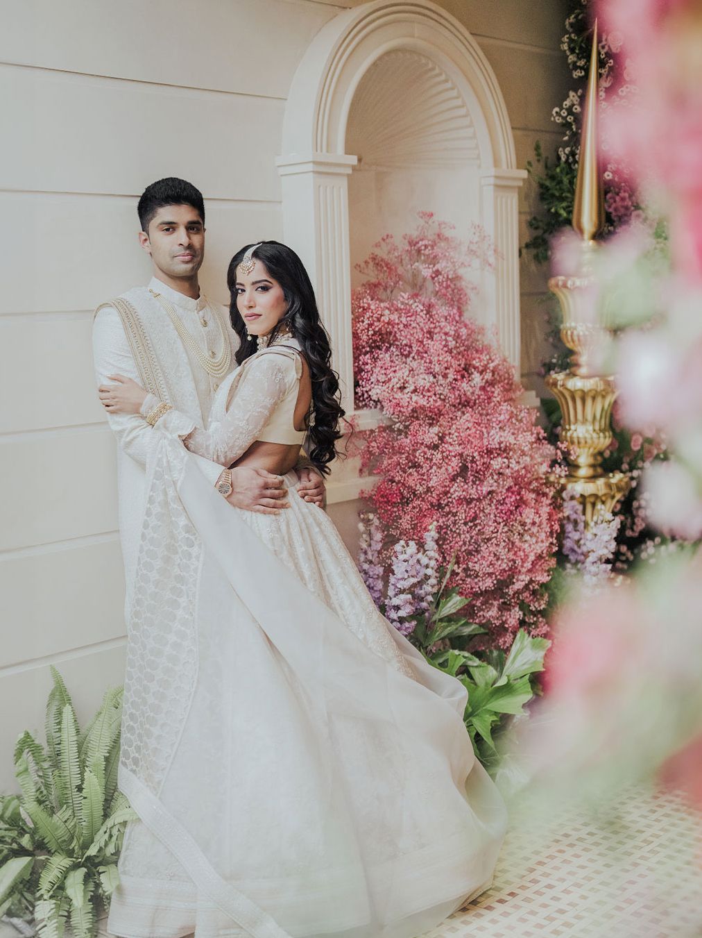 A couple in elegant white Indian wedding attire poses before lush pink floral arrangements in a romantic garden setting.