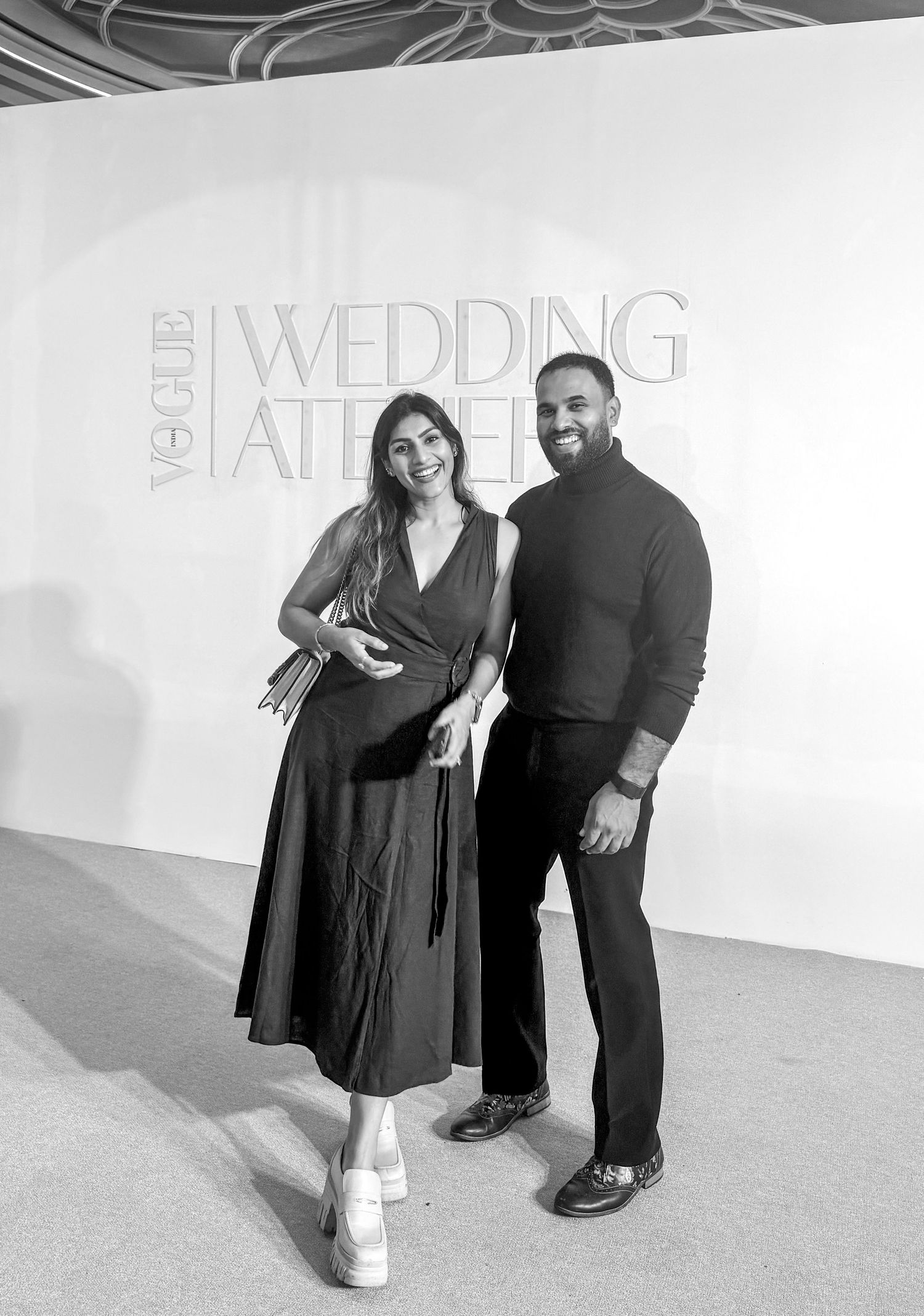 Couple posing at Vogue Wedding Atelier event backdrop, woman in midi dress, man in black turtleneck, black and white photo.