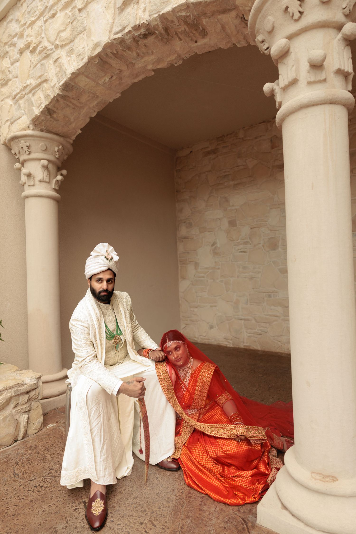 Indian couple in traditional wedding attire posing under ornate stone arch, groom in white sherwani, bride in red saree.