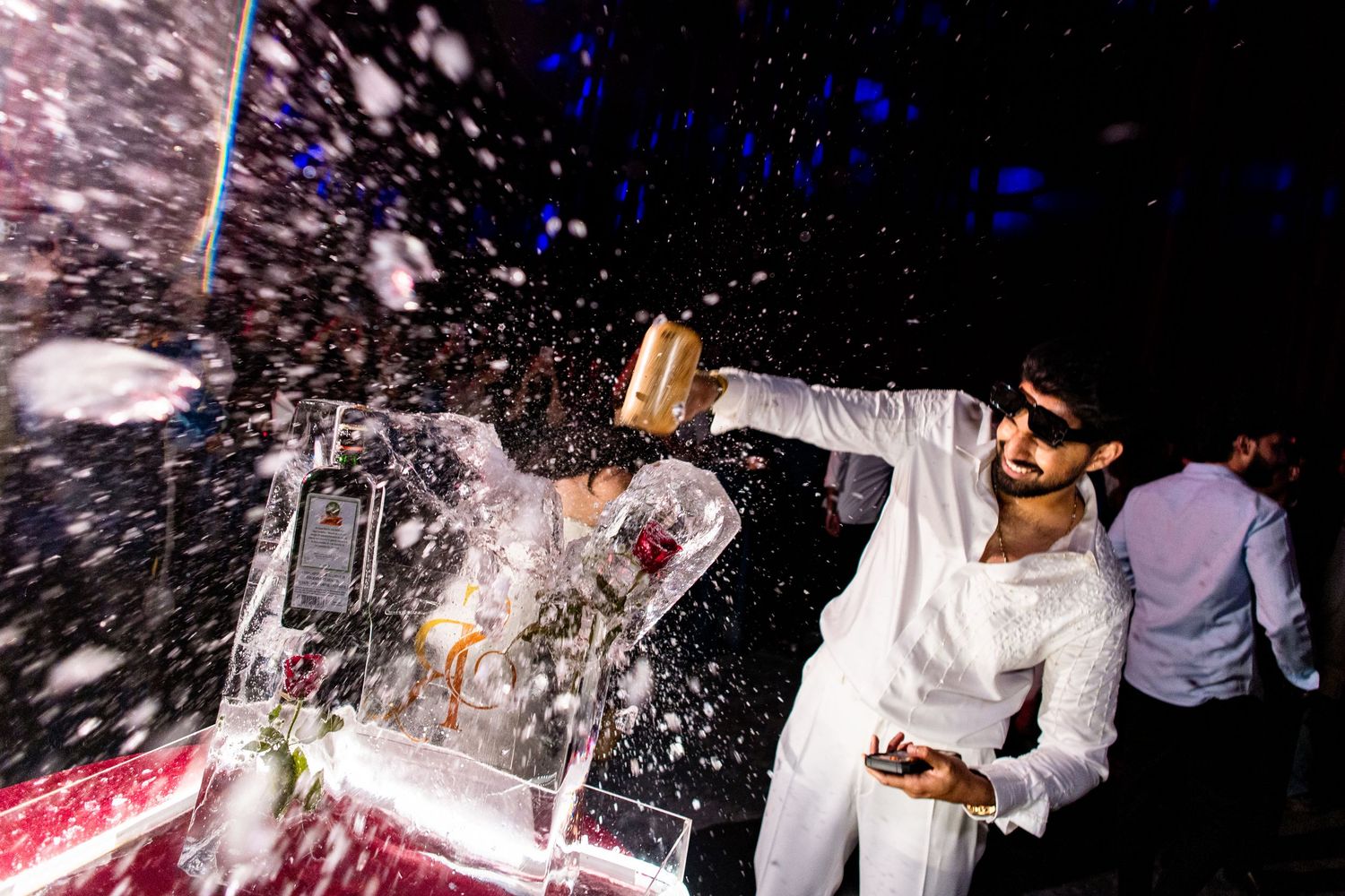 Man in white suit spraying champagne over an ice sculpture at a lively celebration party.