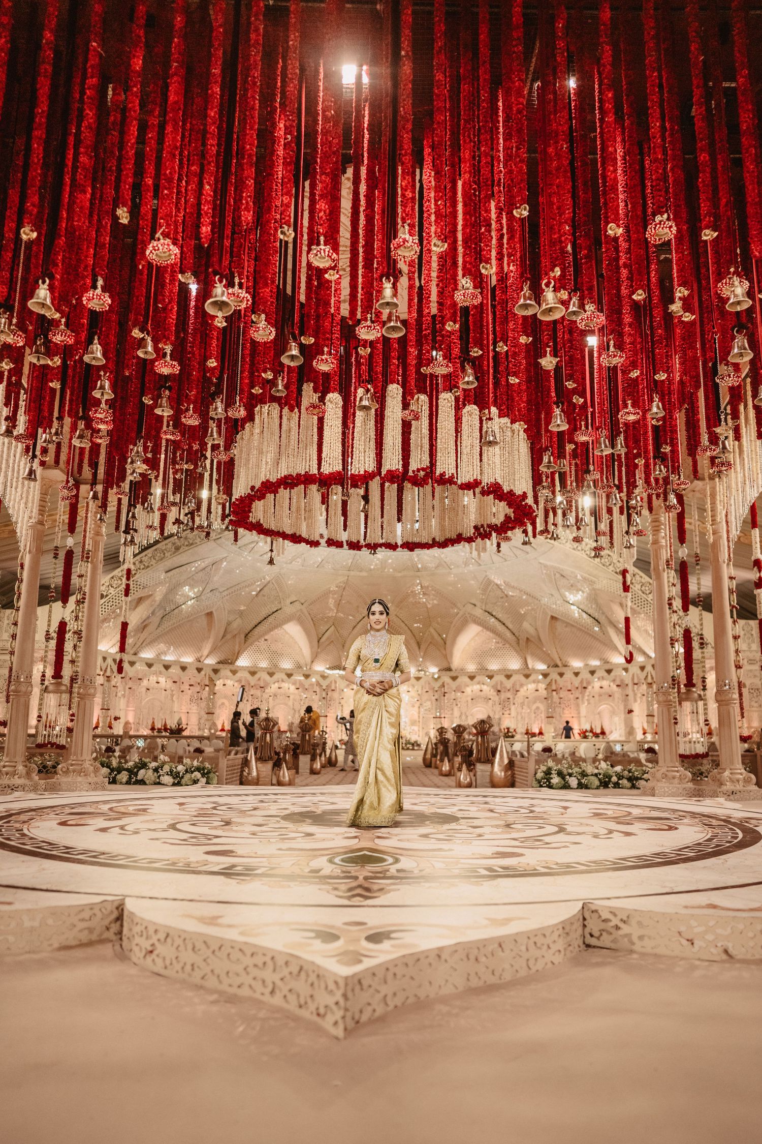 Bride in gold saree stands beneath stunning red floral ceiling installation at luxurious Indian wedding venue.