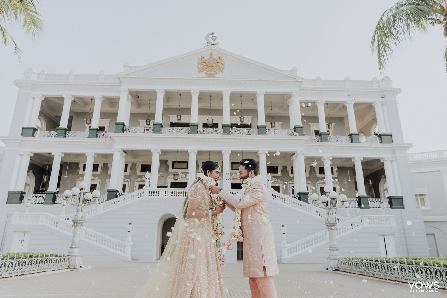 Bride and groom in traditional Indian wedding attire pose before a grand white colonial palace venue.