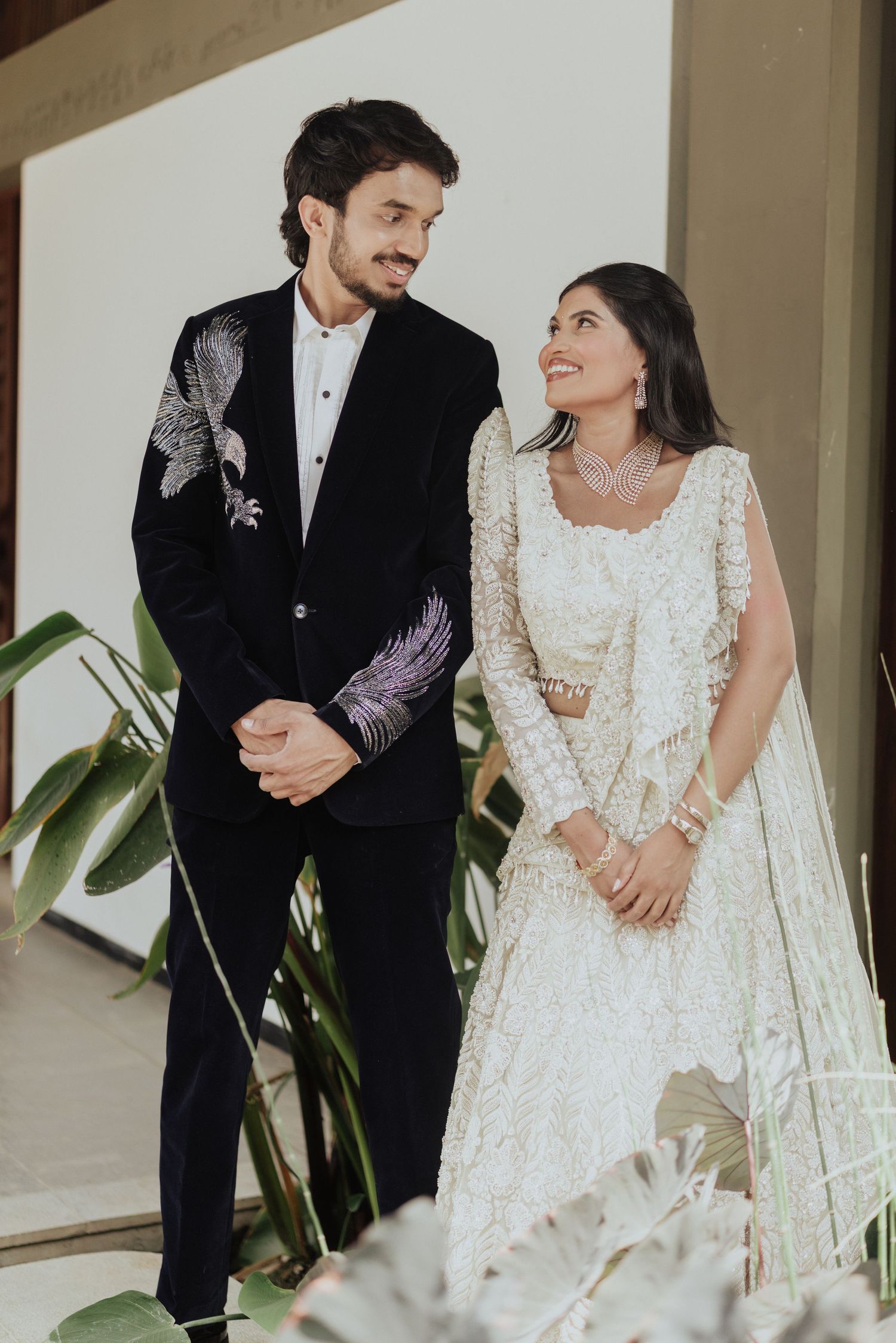 Indian couple in elegant wedding attire smile at each other, bride in white lehenga, groom in black embroidered suit.