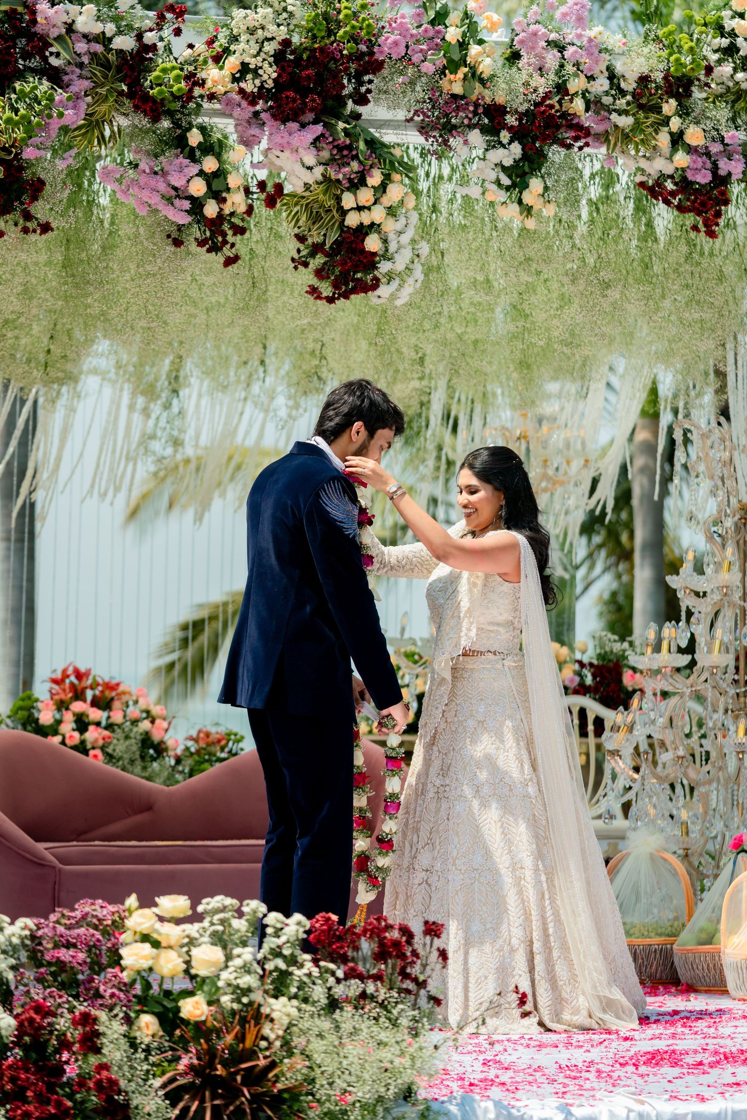 Bride places garland on groom under lush floral canopy with hanging greenery and colorful blooms at Indian wedding.