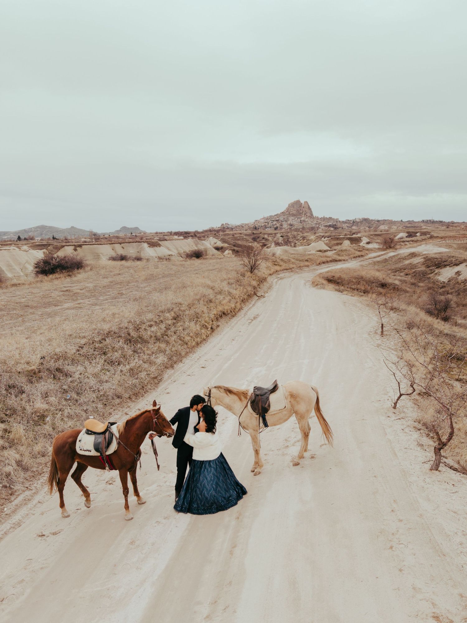 Couple in black attire with two saddled horses on a sandy desert road in Cappadocia, Turkey.
