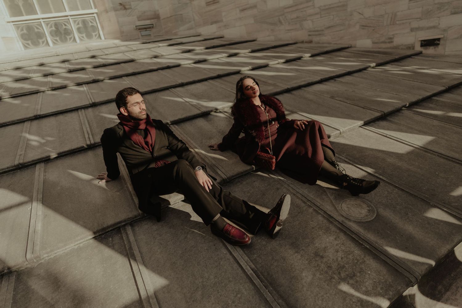 A stylish couple in dark red outfits sits relaxed on the geometric tiled rooftop of a cathedral.