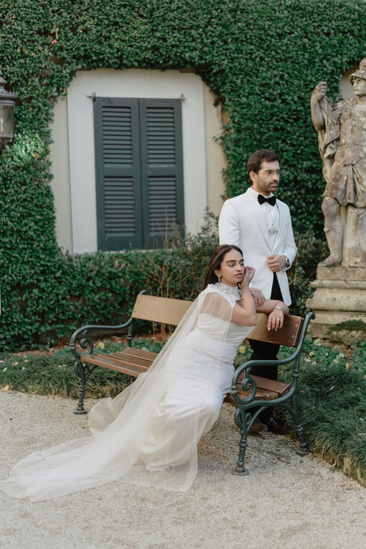 Bride in white gown sits on bench while groom in white tuxedo stands beside her at ivy-covered villa.