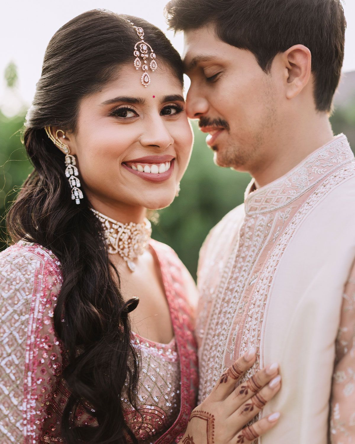 Indian bride and groom in traditional wedding attire smiling closely together, bride wearing jewelry and mehndi.