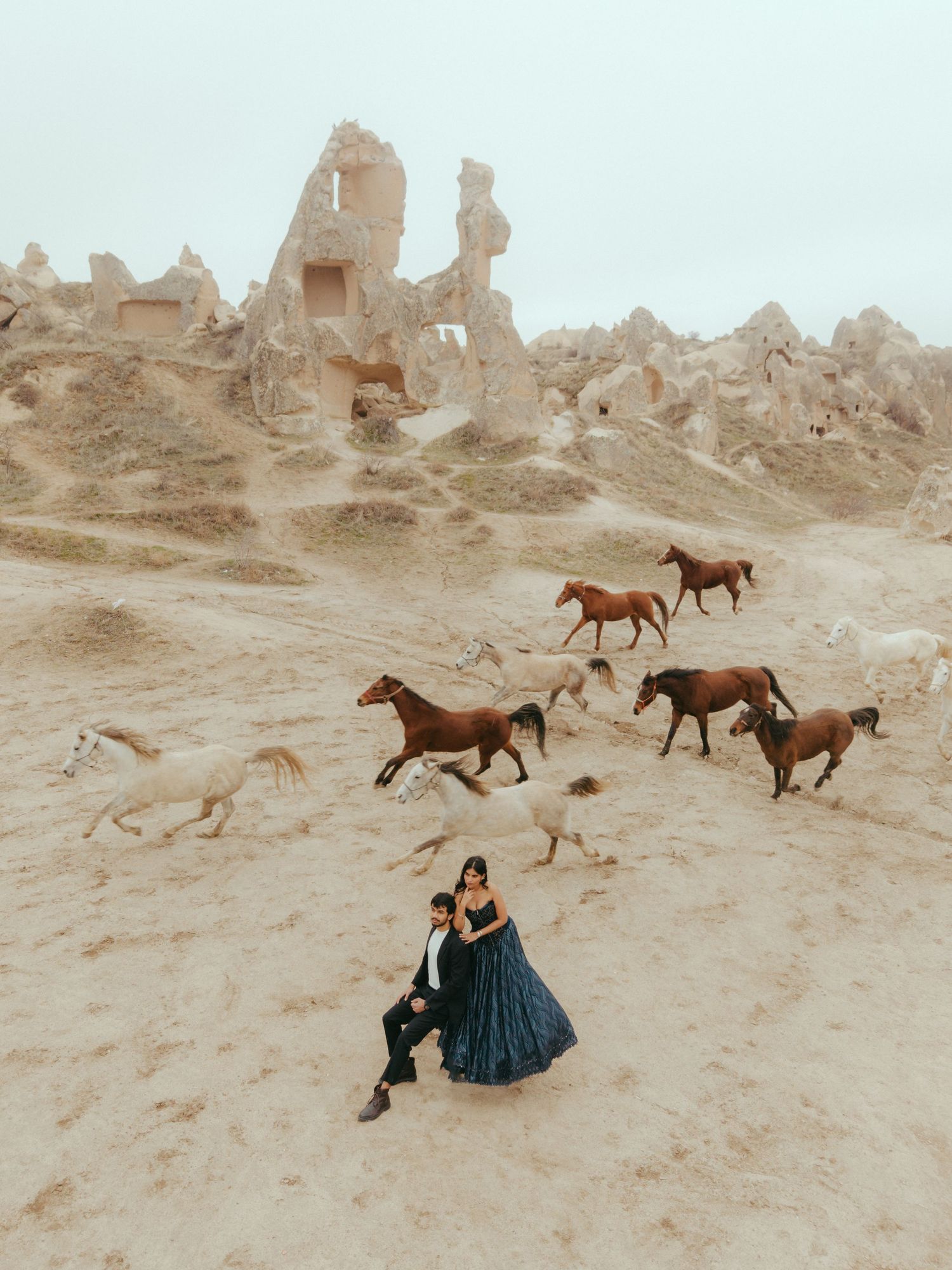 Couple sitting together surrounded by wild horses galloping near ancient rock ruins in Cappadocia.
