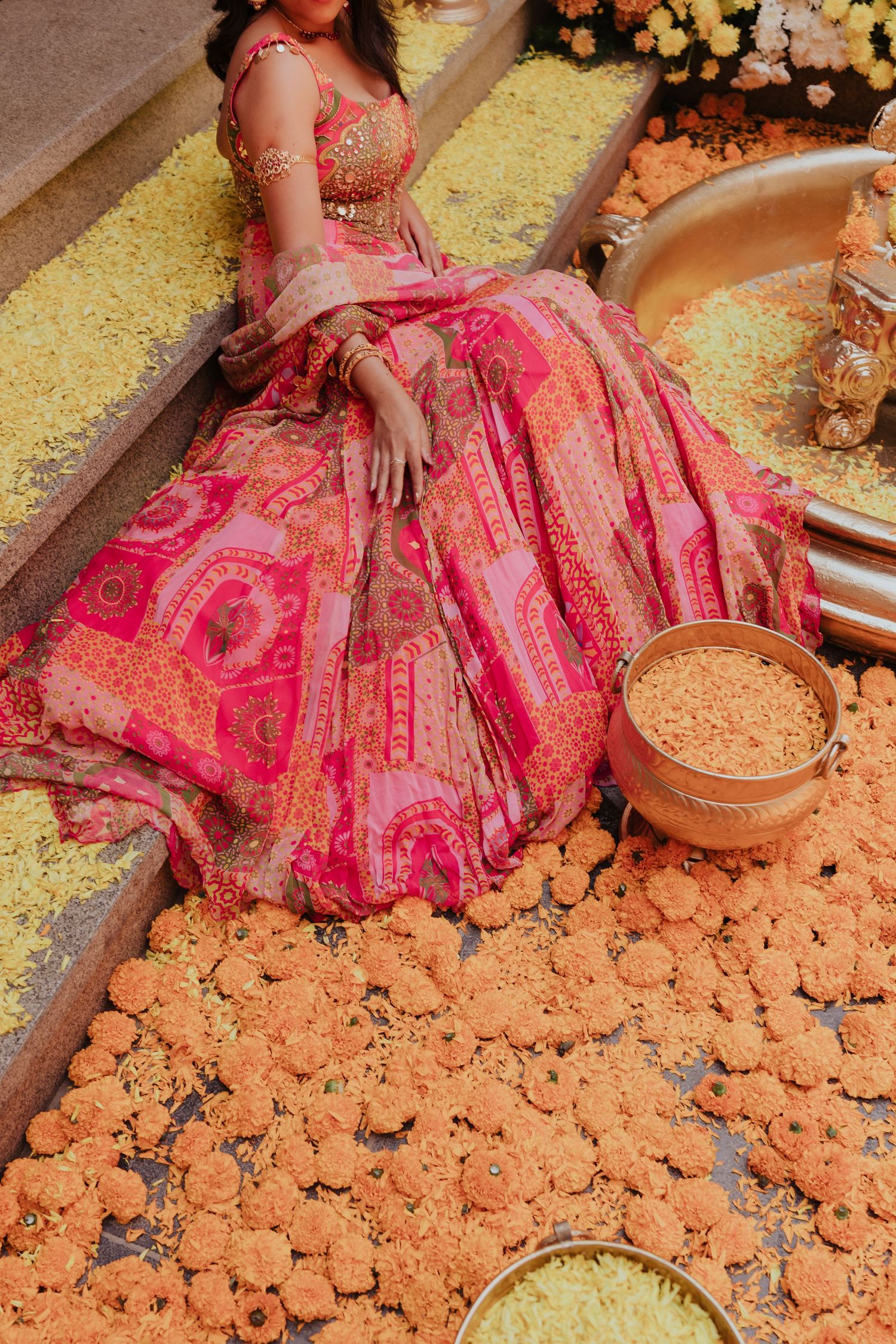 Bride in vibrant pink printed lehenga seated among marigold flowers during a traditional Indian wedding ceremony.