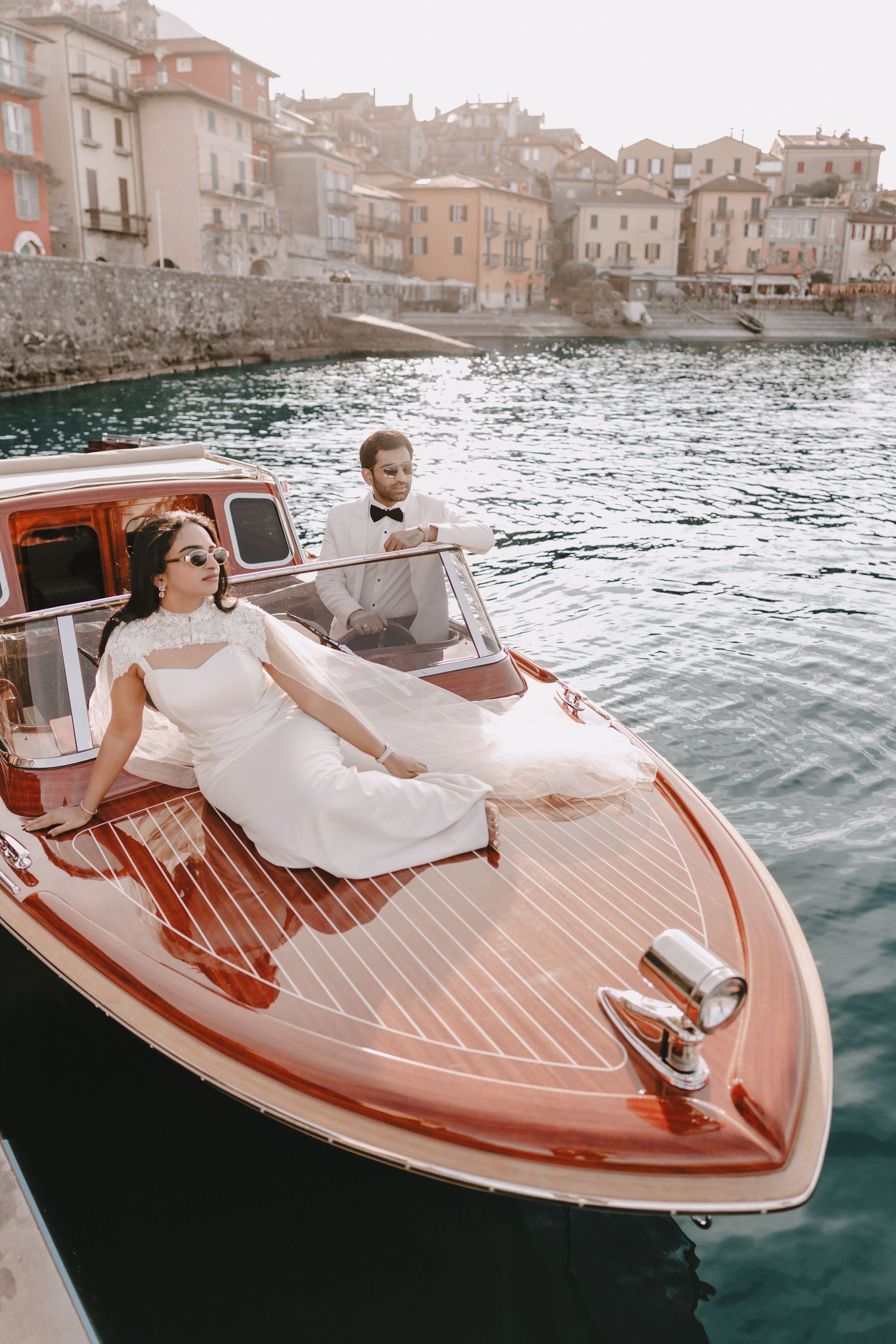 Bride and groom posing on a classic wooden speedboat with colorful Italian village behind them.