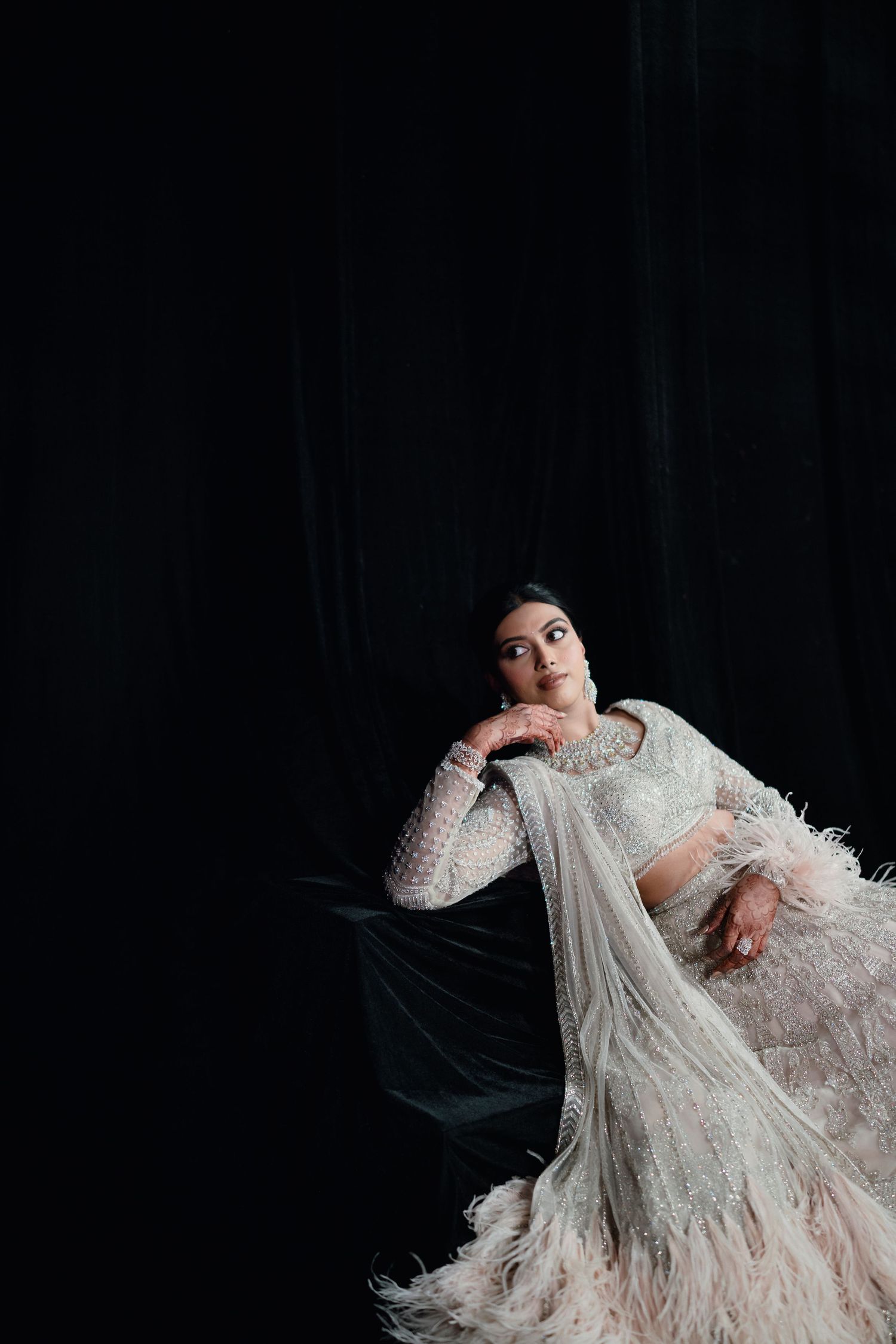 Woman in white feathered lehenga poses gracefully against dark backdrop in elegant bridal fashion photography.