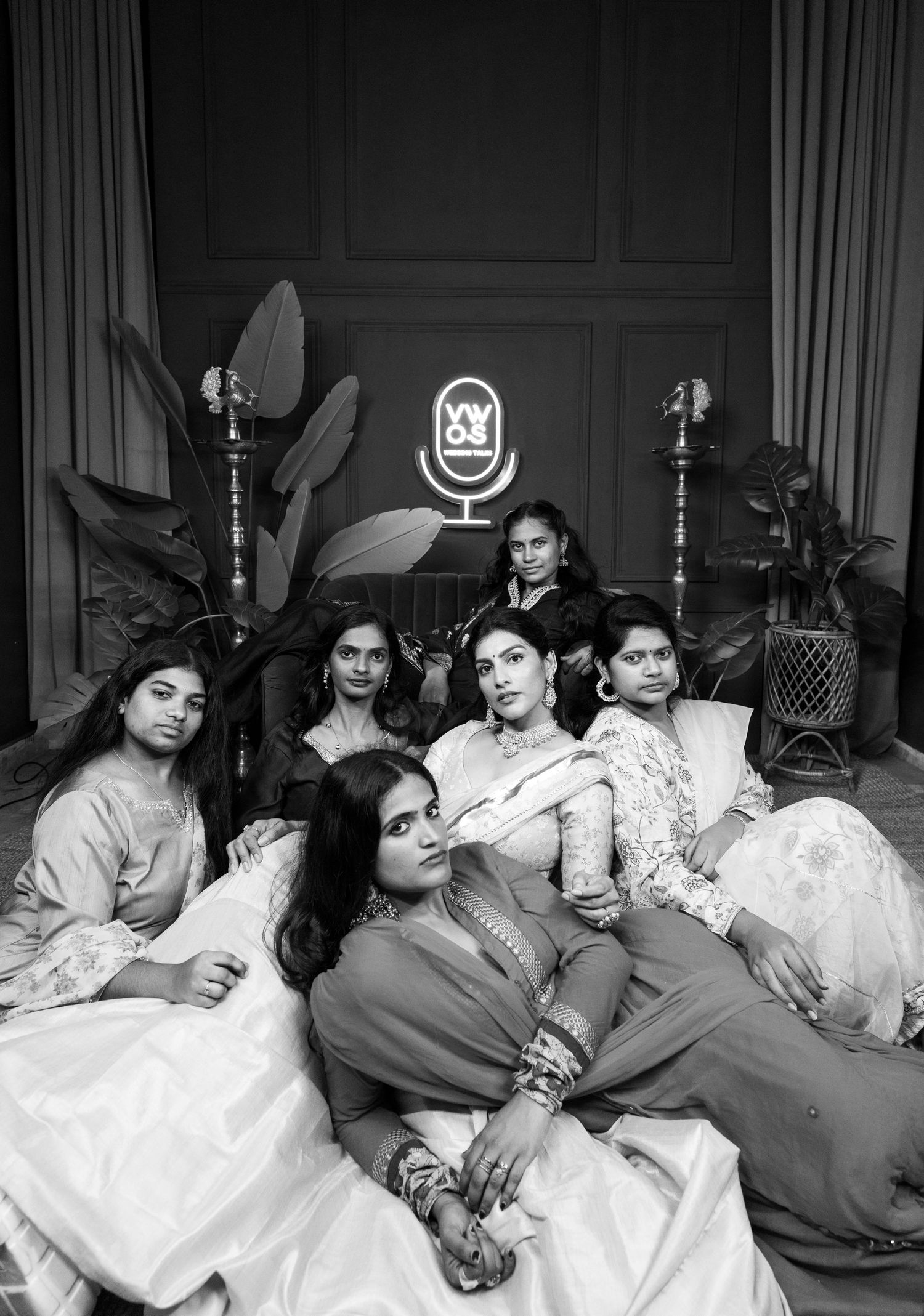 Black and white photo of six women in traditional Indian attire posing elegantly in a stylish studio setting.
