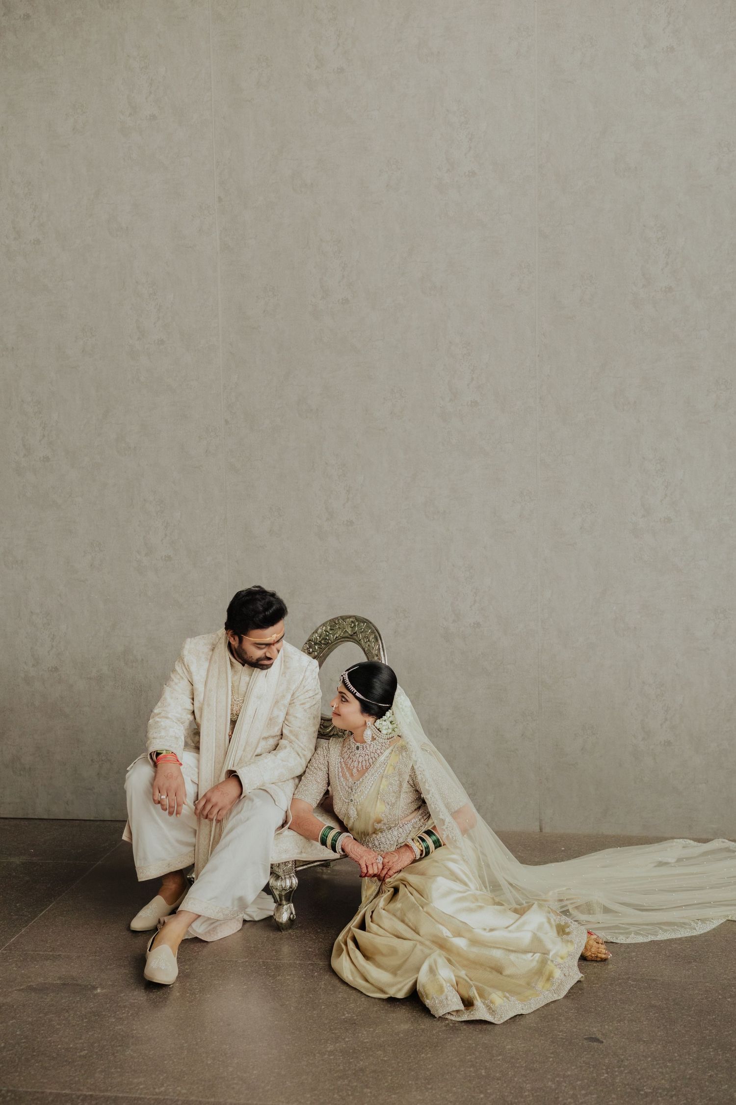 Indian bride and groom in ivory wedding attire sitting together on floor against a neutral textured wall.