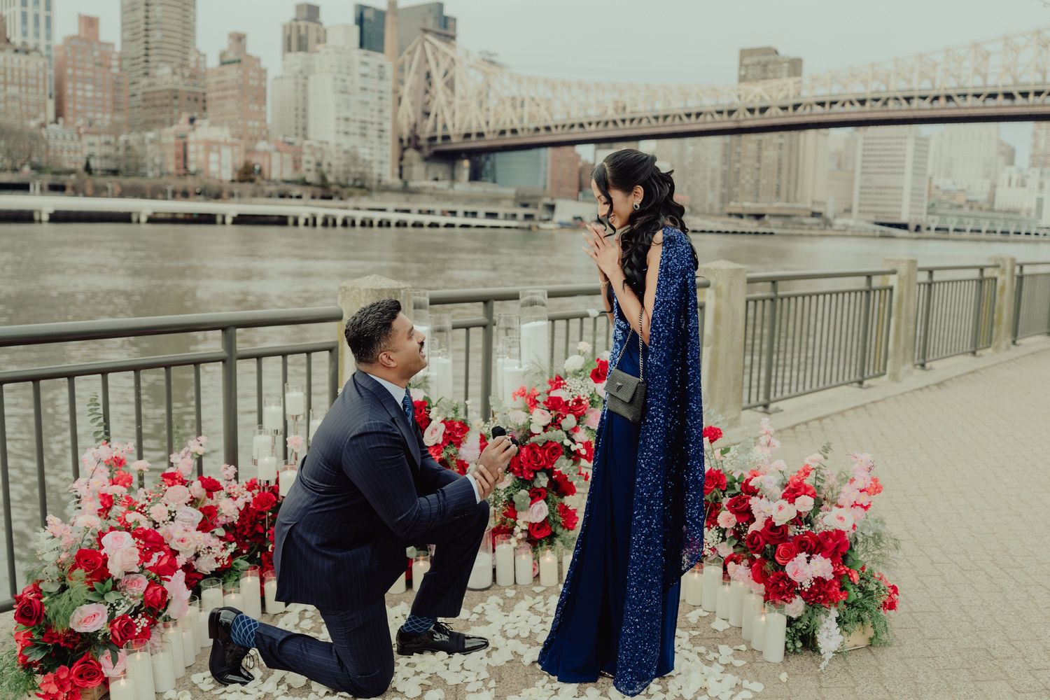 Man proposes to woman in blue dress by NYC waterfront with red and pink floral arrangements and bridge backdrop.