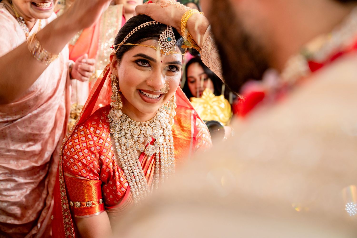 Radiant Indian bride in red lehenga and gold jewelry smiling during traditional Hindu wedding ceremony.