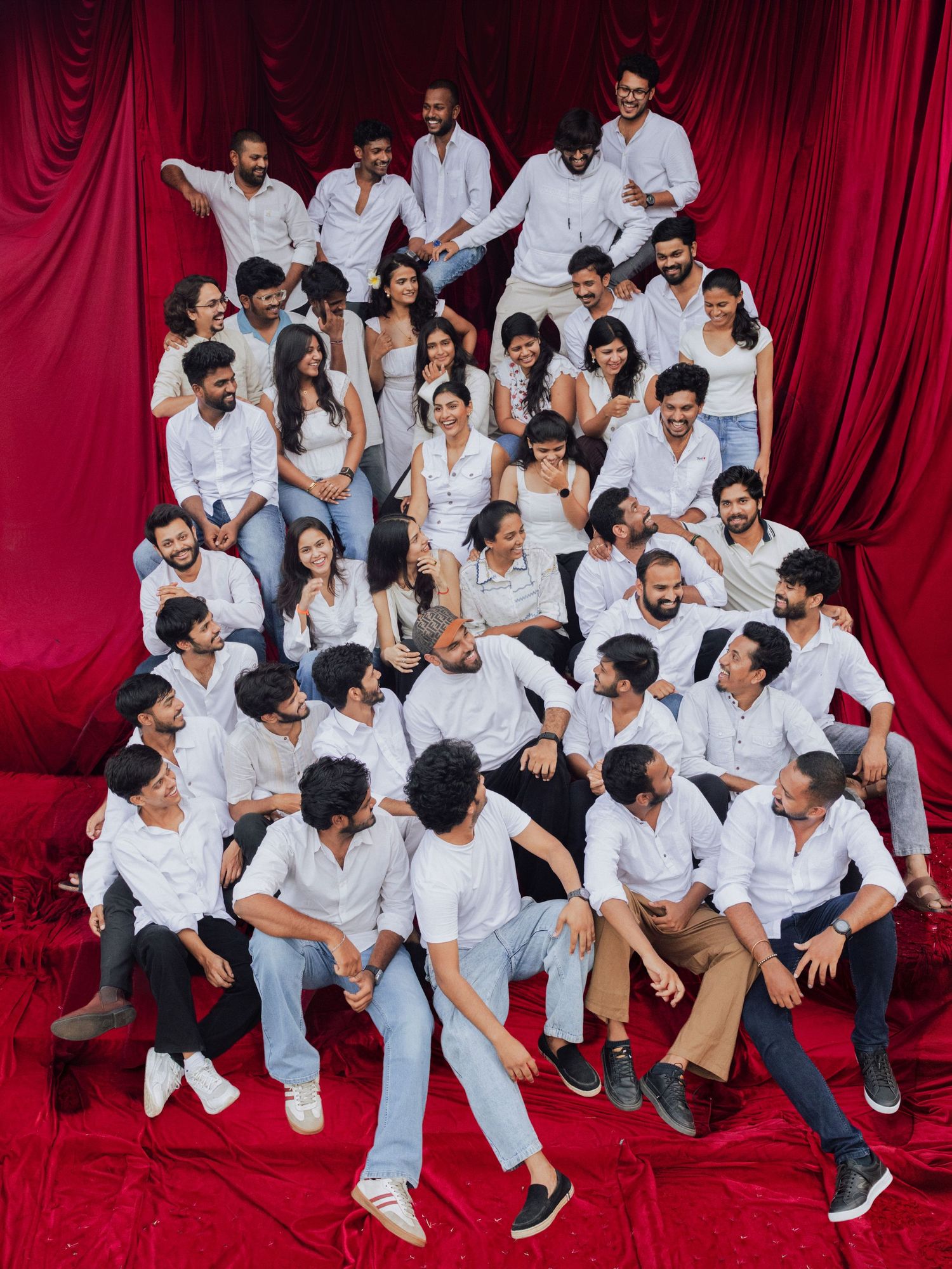 Large group of young adults in white shirts and jeans posing together against a red curtain backdrop.