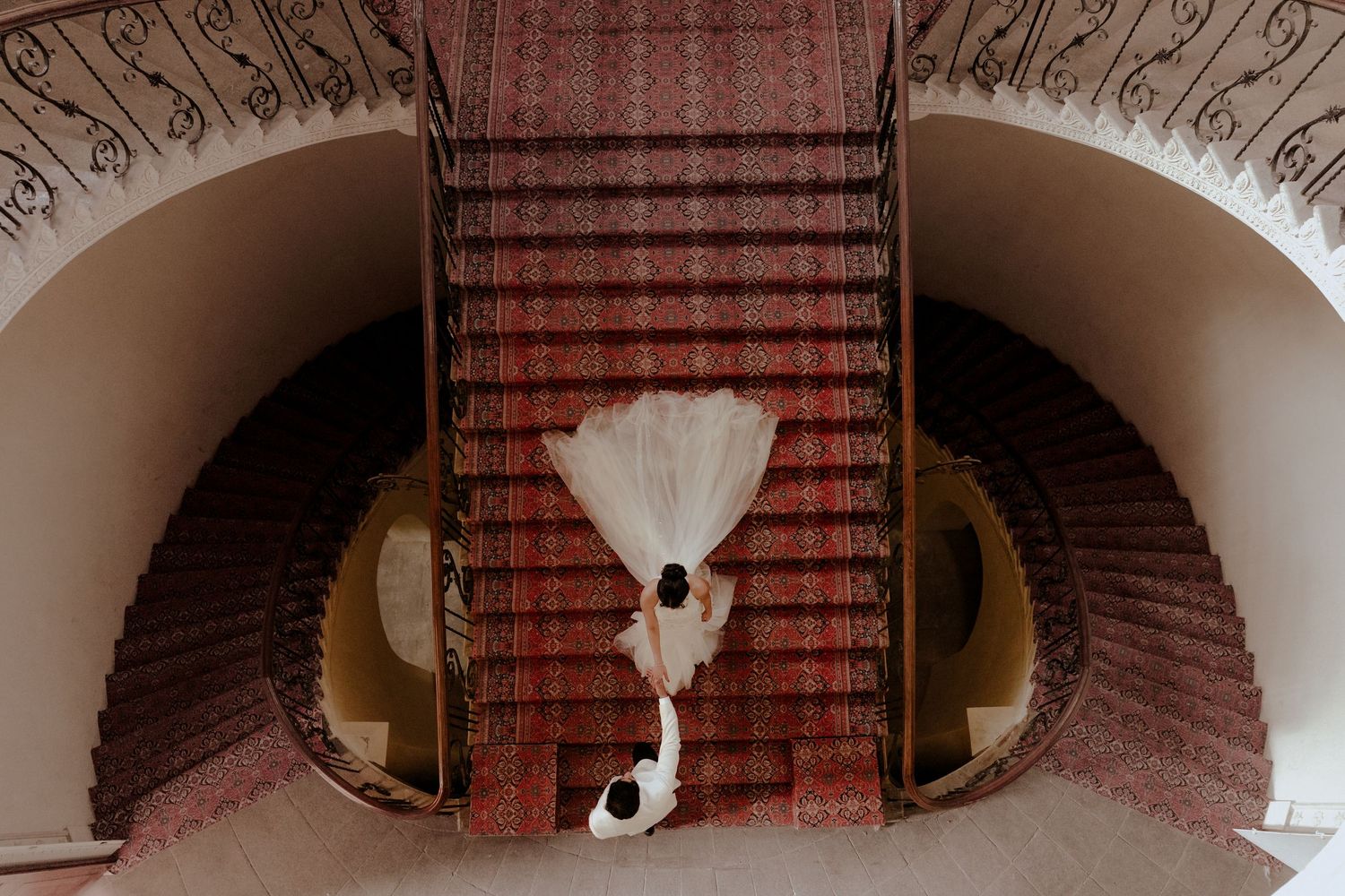 Overhead view of a bride in a white gown with a long train standing on a grand red-carpeted curved staircase.