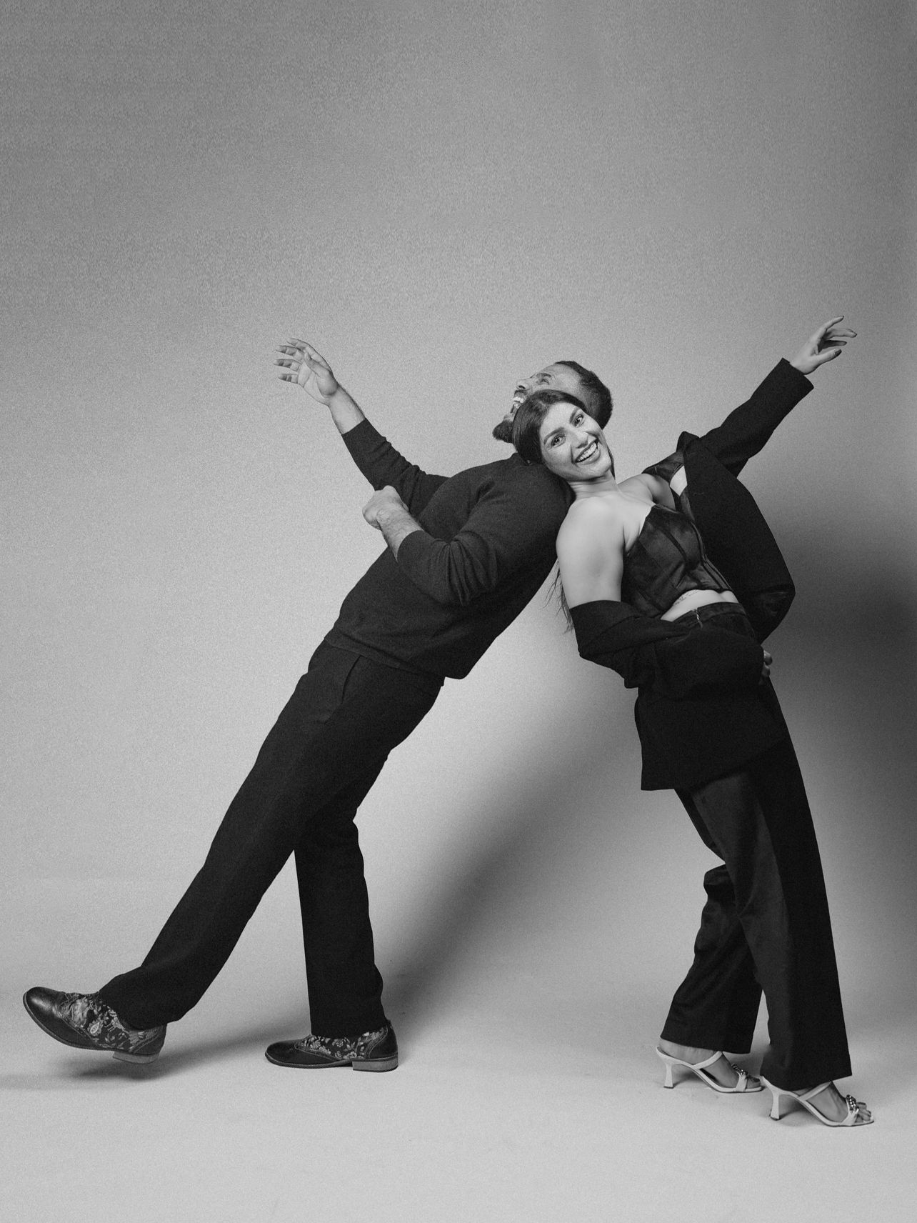 Two dancers in black outfits strike dynamic poses in a black and white studio photo.