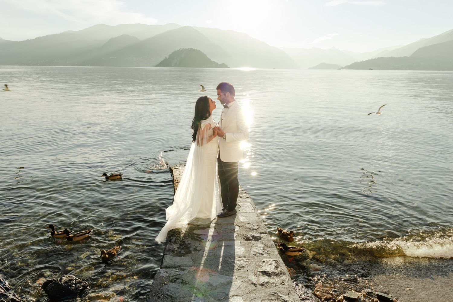 Newlyweds embrace on sunlit lakeside pier surrounded by ducks with misty mountains behind them.