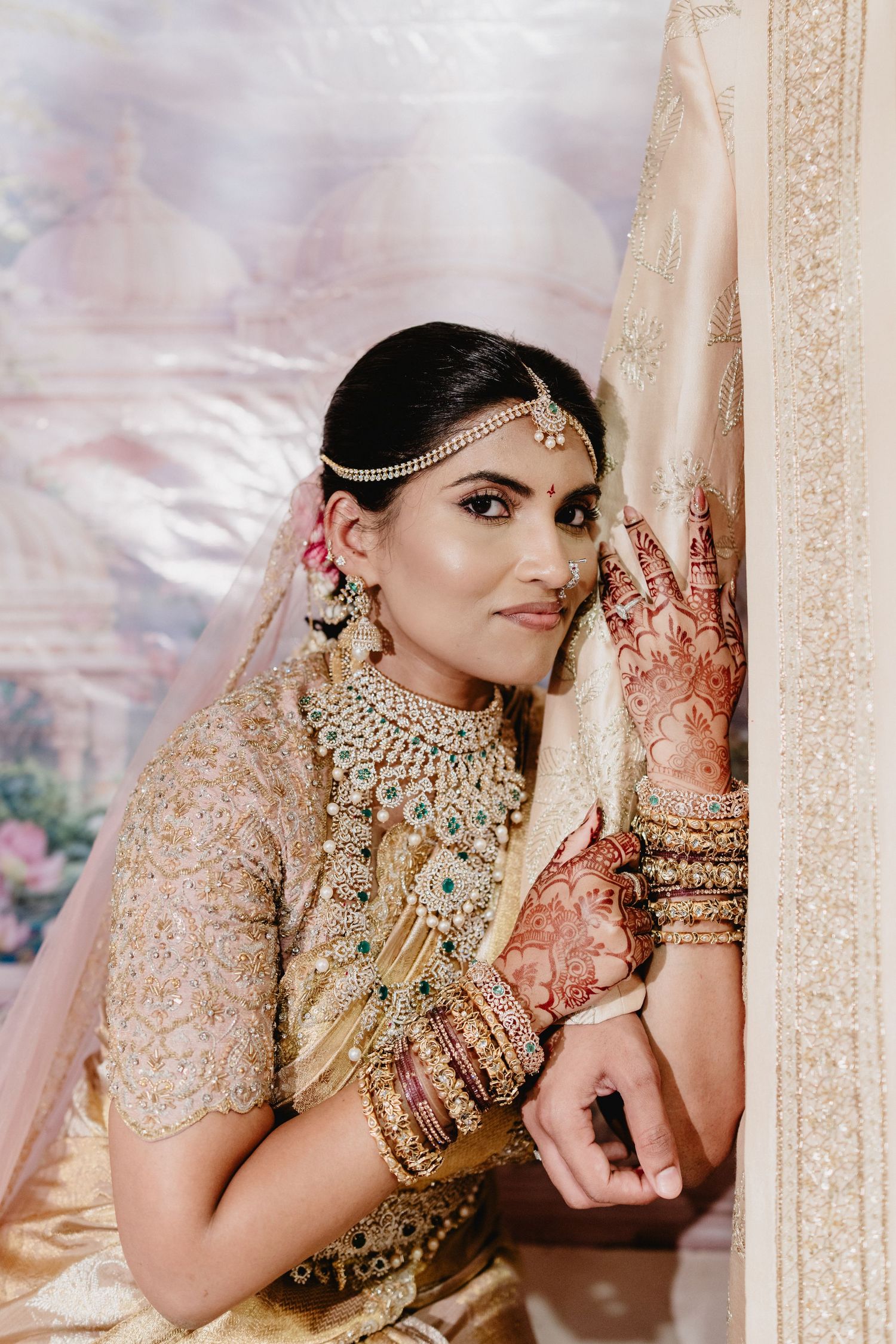Indian bride in gold lehenga with intricate henna, jeweled necklace, maang tikka, and bangles poses gracefully in bridal attire.