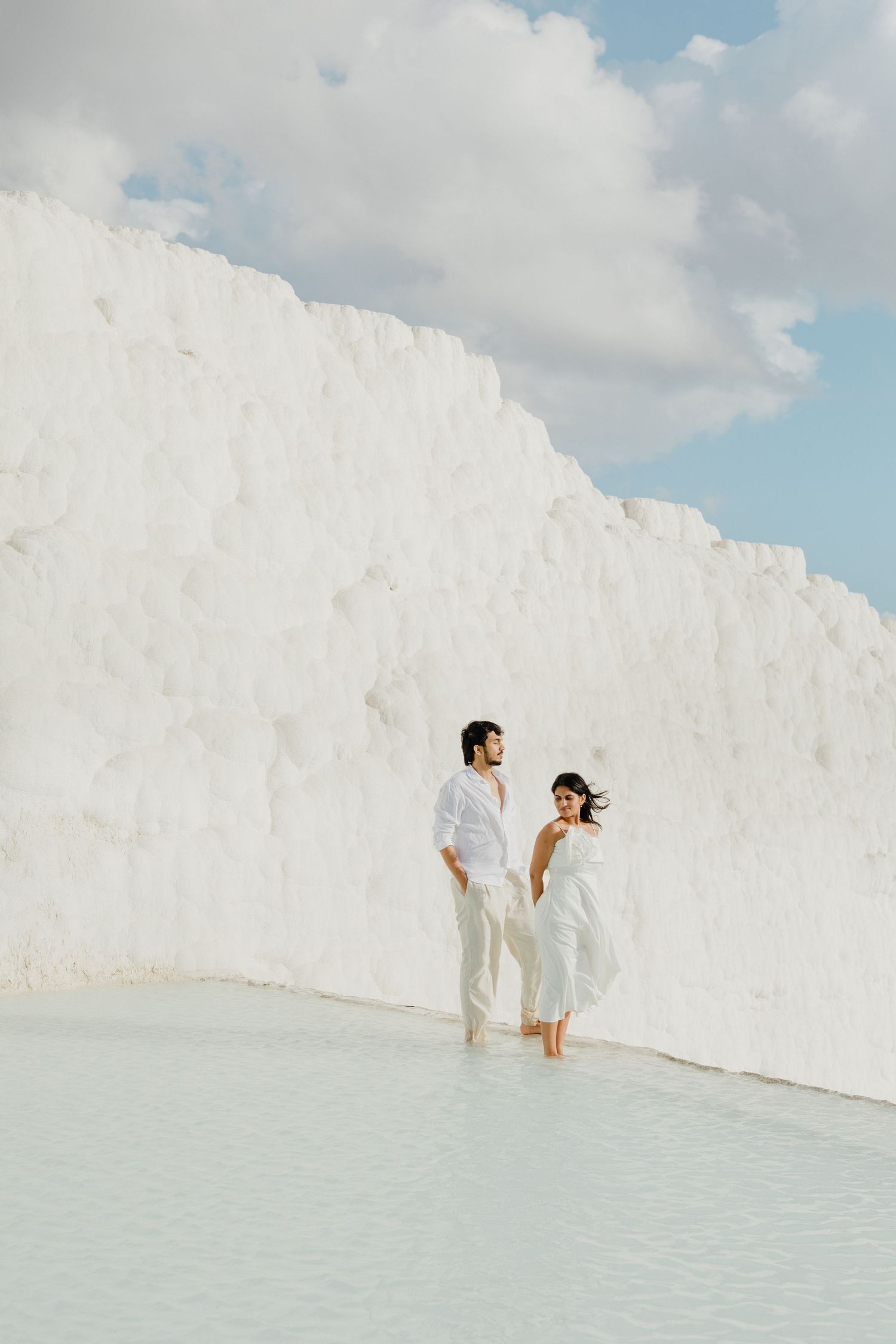 Couple in white outfits walking through Pamukkale travertine pools with stunning white calcium terraces backdrop.