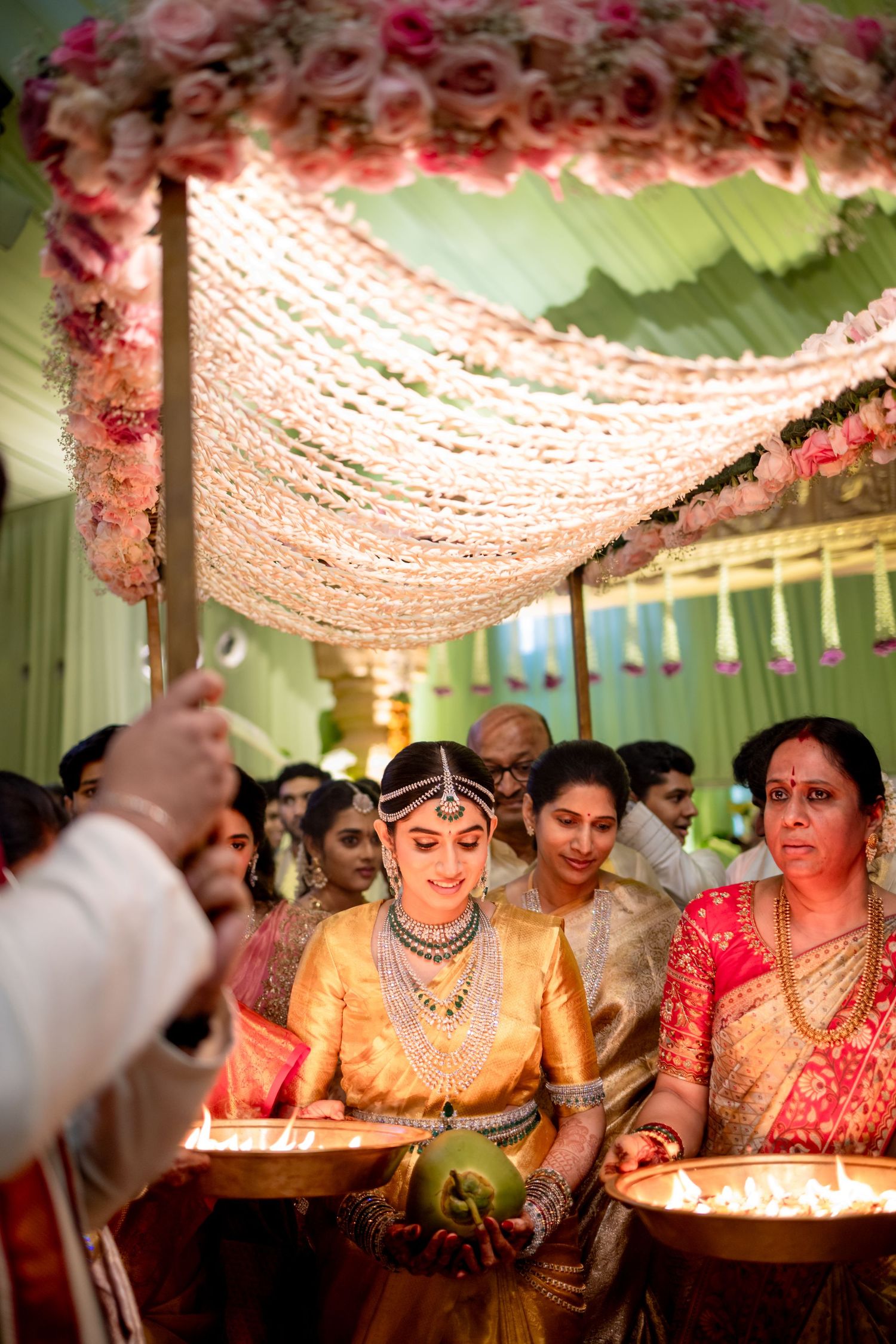 South Indian bride in golden saree walks under floral mandap holding coconut during traditional wedding ceremony.