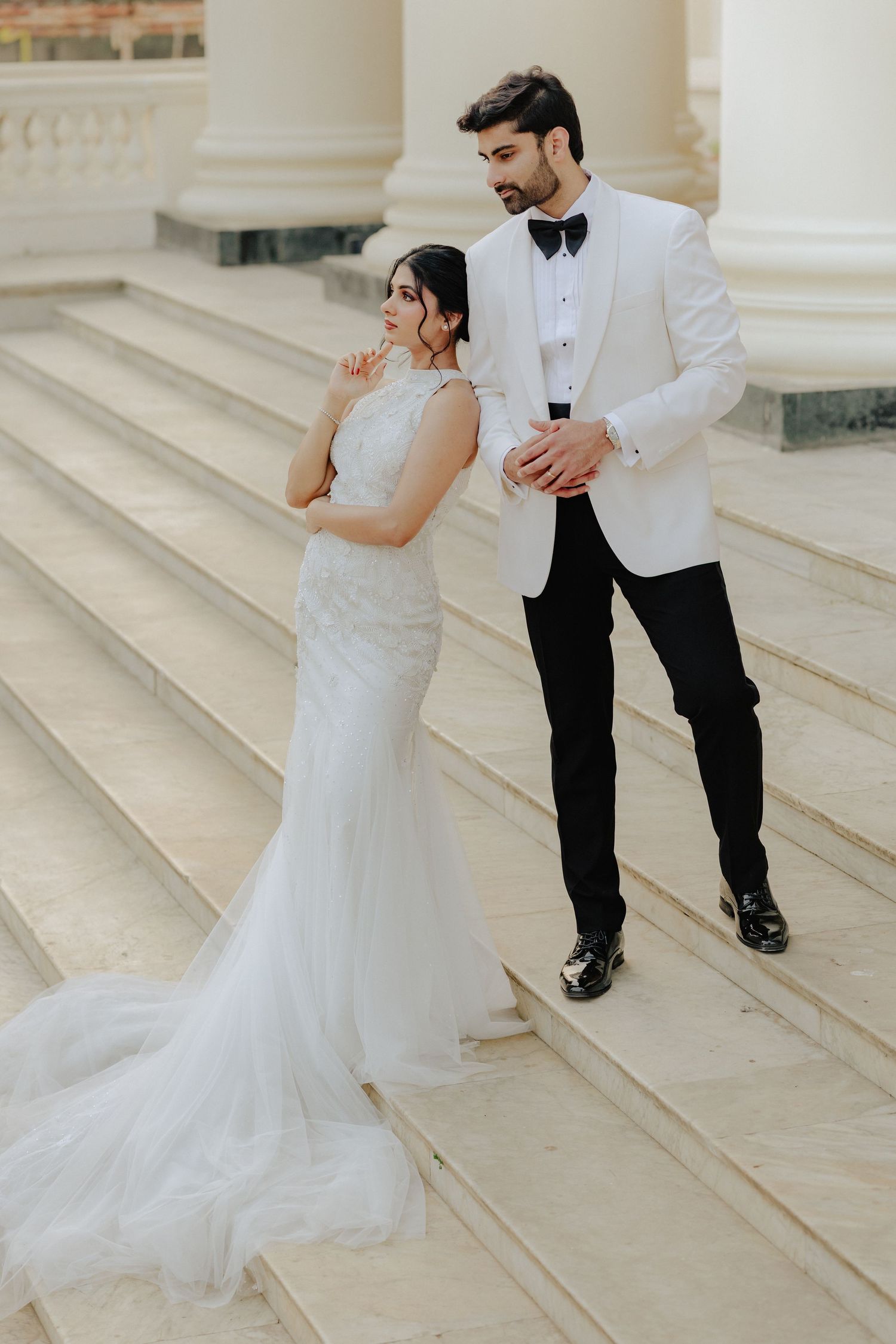 Bride in white mermaid gown stands beside groom in white tuxedo on stone steps.
