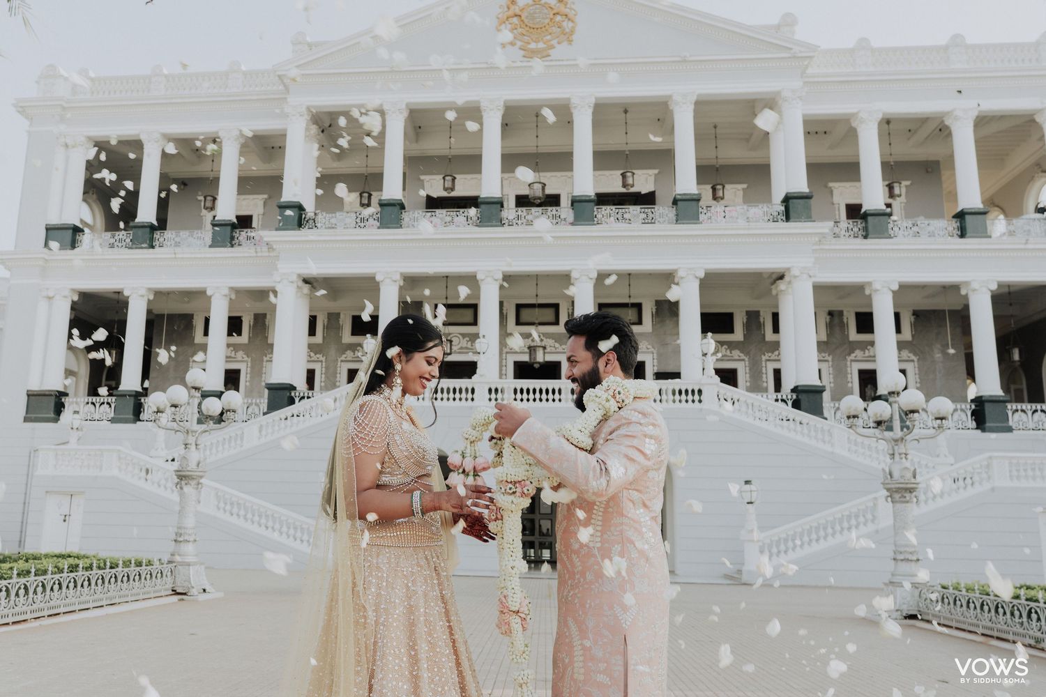 Couple in traditional Indian wedding attire exchange vows outdoors at elegant white venue with flower petals falling.