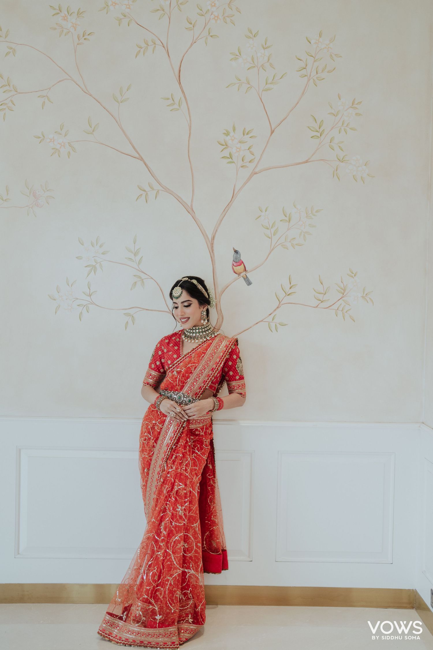Bride in red embroidered saree with gold jewelry poses elegantly against a wall with delicate tree mural.