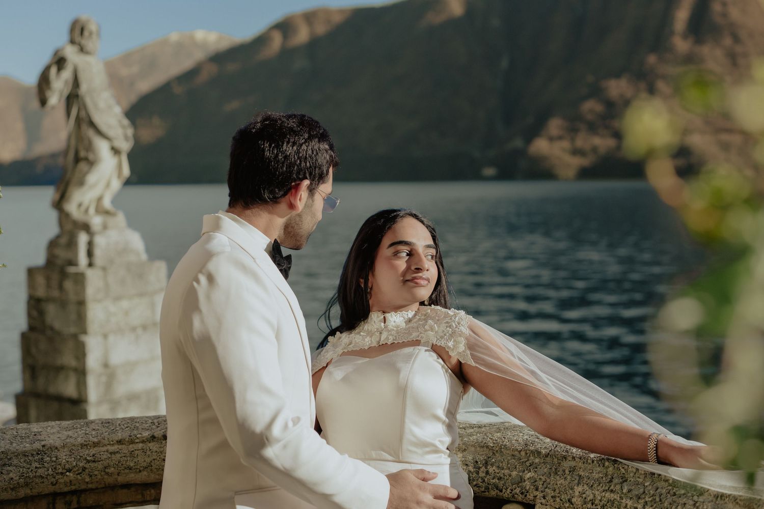 Couple in wedding attire embracing on a lakeside terrace with mountain backdrop at Lake Como, Italy.