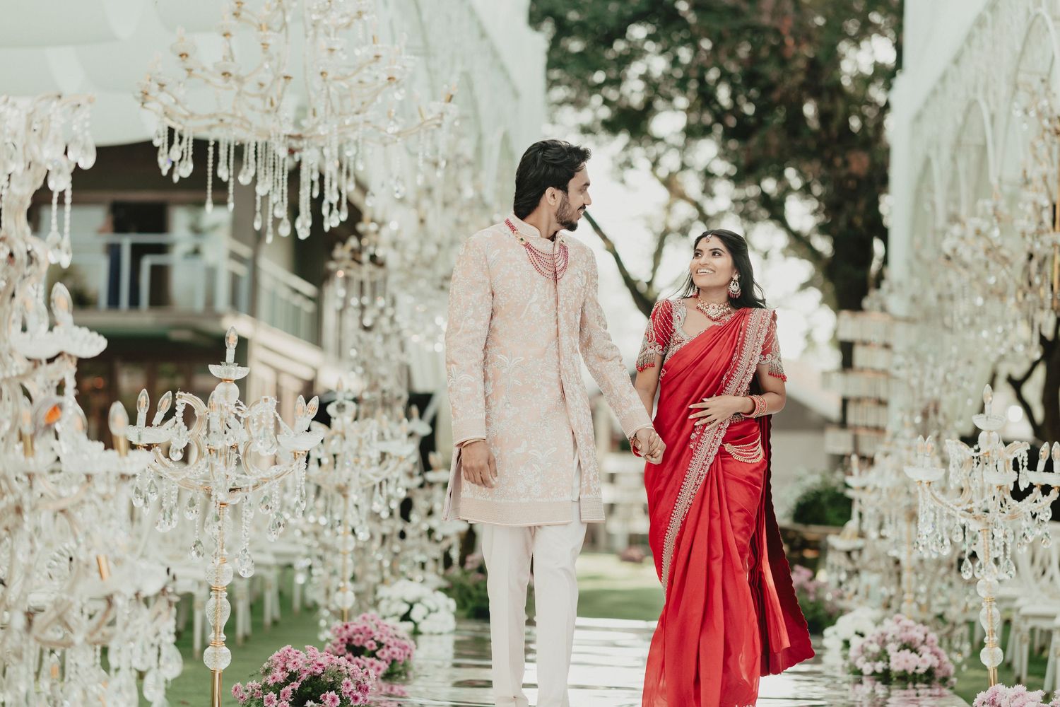 Indian wedding couple in red saree and cream sherwani posing among crystal chandelier decorations.