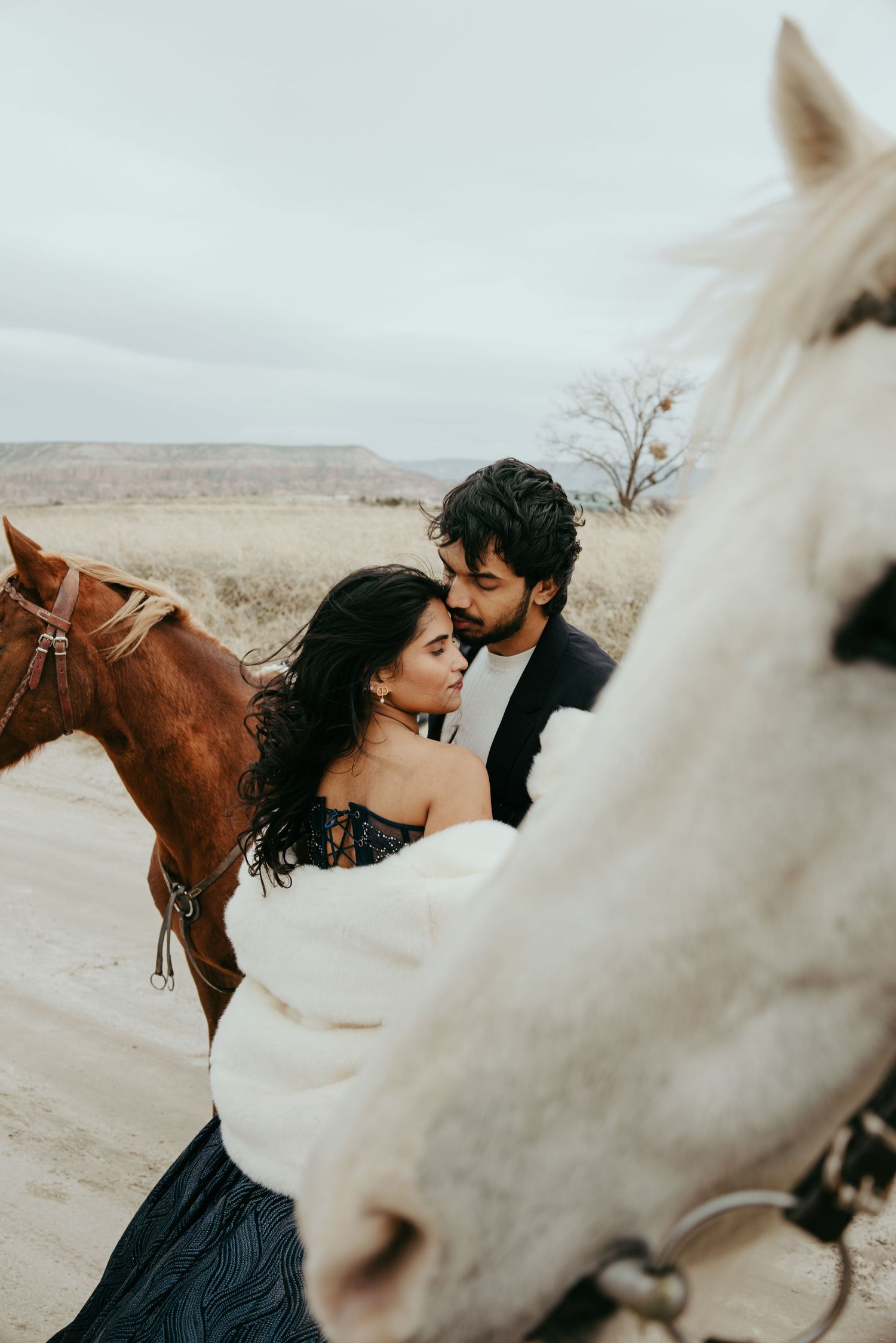 Romantic couple sharing an intimate moment between two horses in a scenic desert landscape.