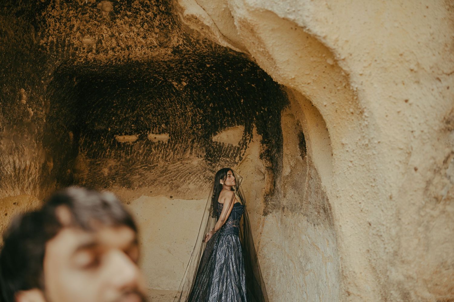 Woman in black gown standing in ancient cave dwelling in Cappadocia, Turkey, with man in foreground.
