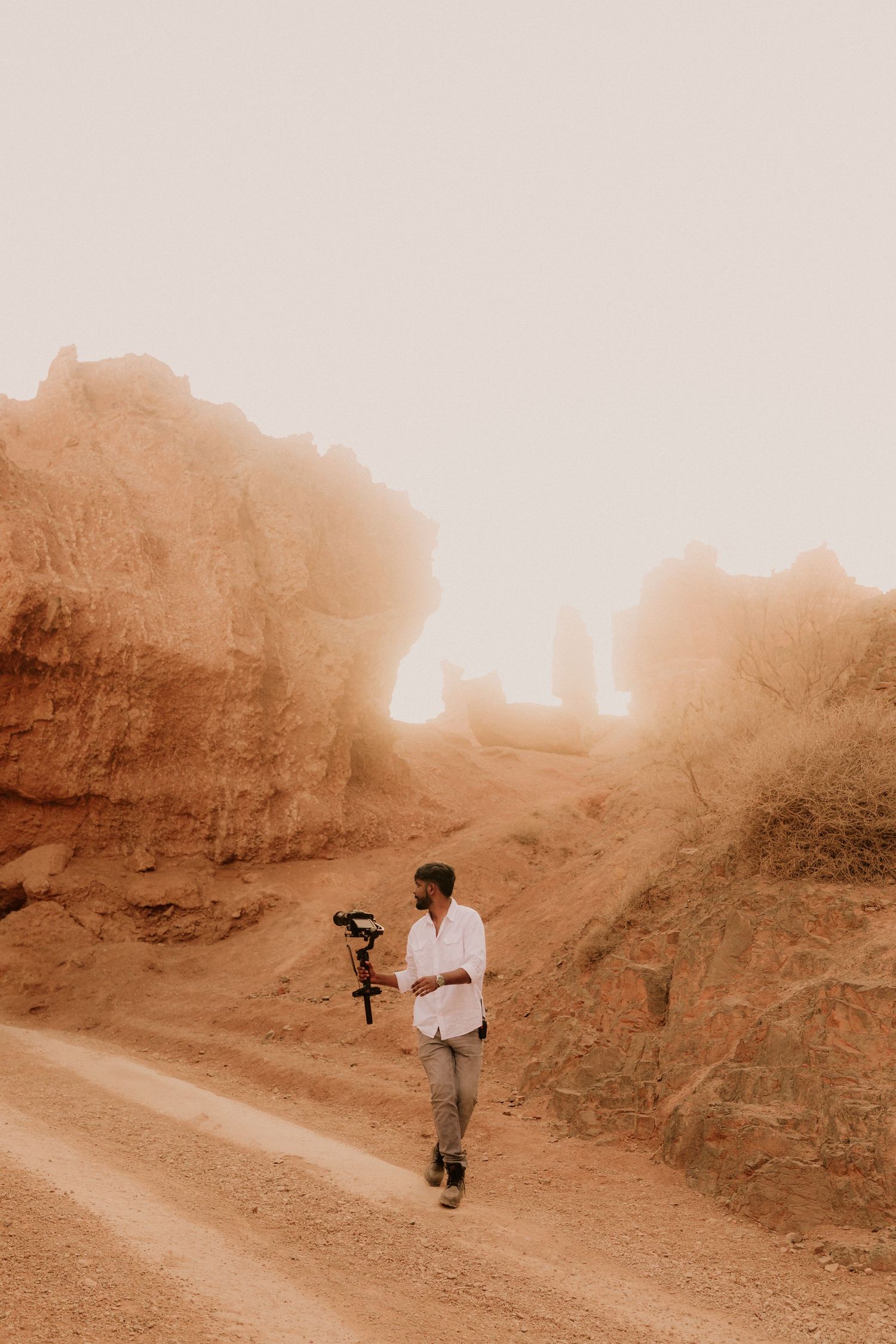 A videographer walks through a misty desert canyon holding a camera stabilizer rig among red rock formations.