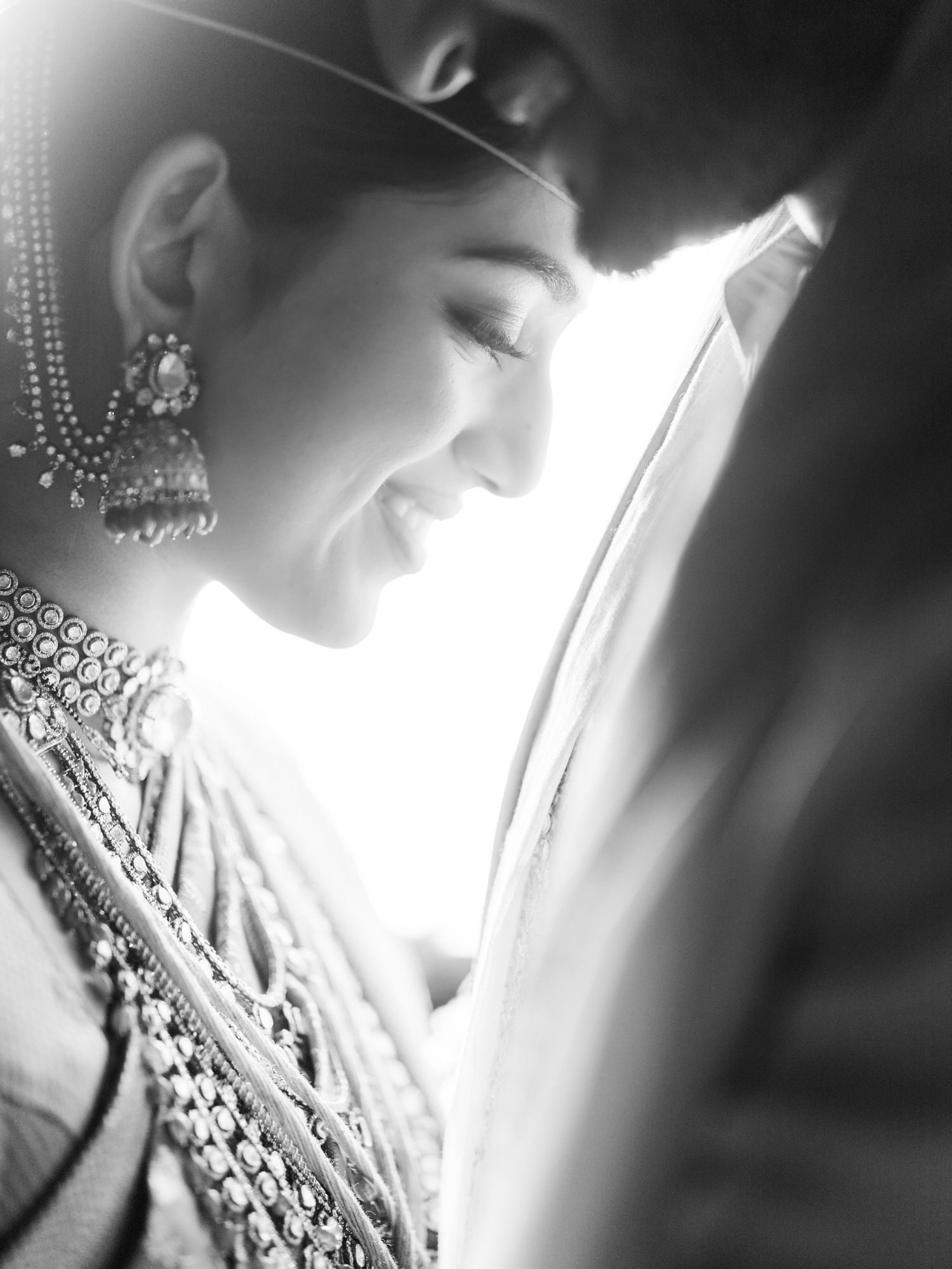 Black and white photo of a smiling Indian bride in traditional jewelry and attire, sharing an intimate moment with her groom.