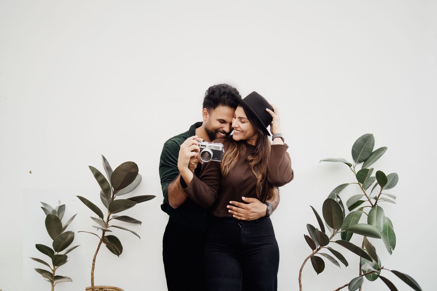 A joyful couple sharing a laugh while holding a vintage camera, surrounded by green plants against a white background.