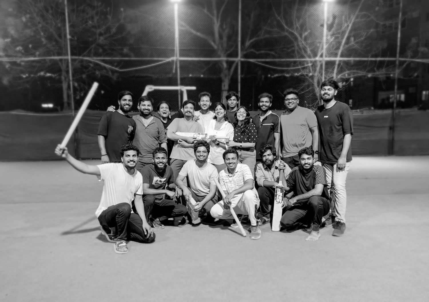 Black and white photo of a group of men posing on a cricket field at night, some holding bats.