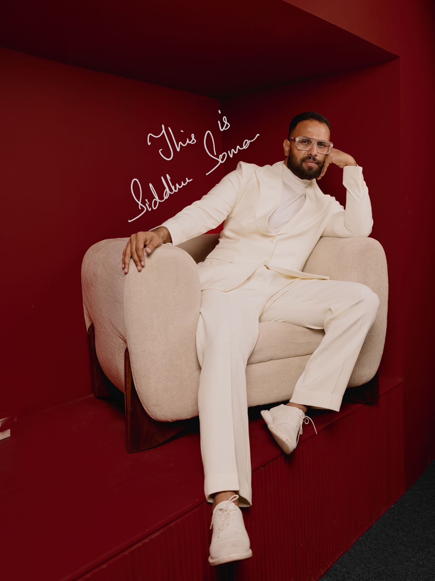 Man in white suit seated on beige chair against red wall with cursive text overlay.