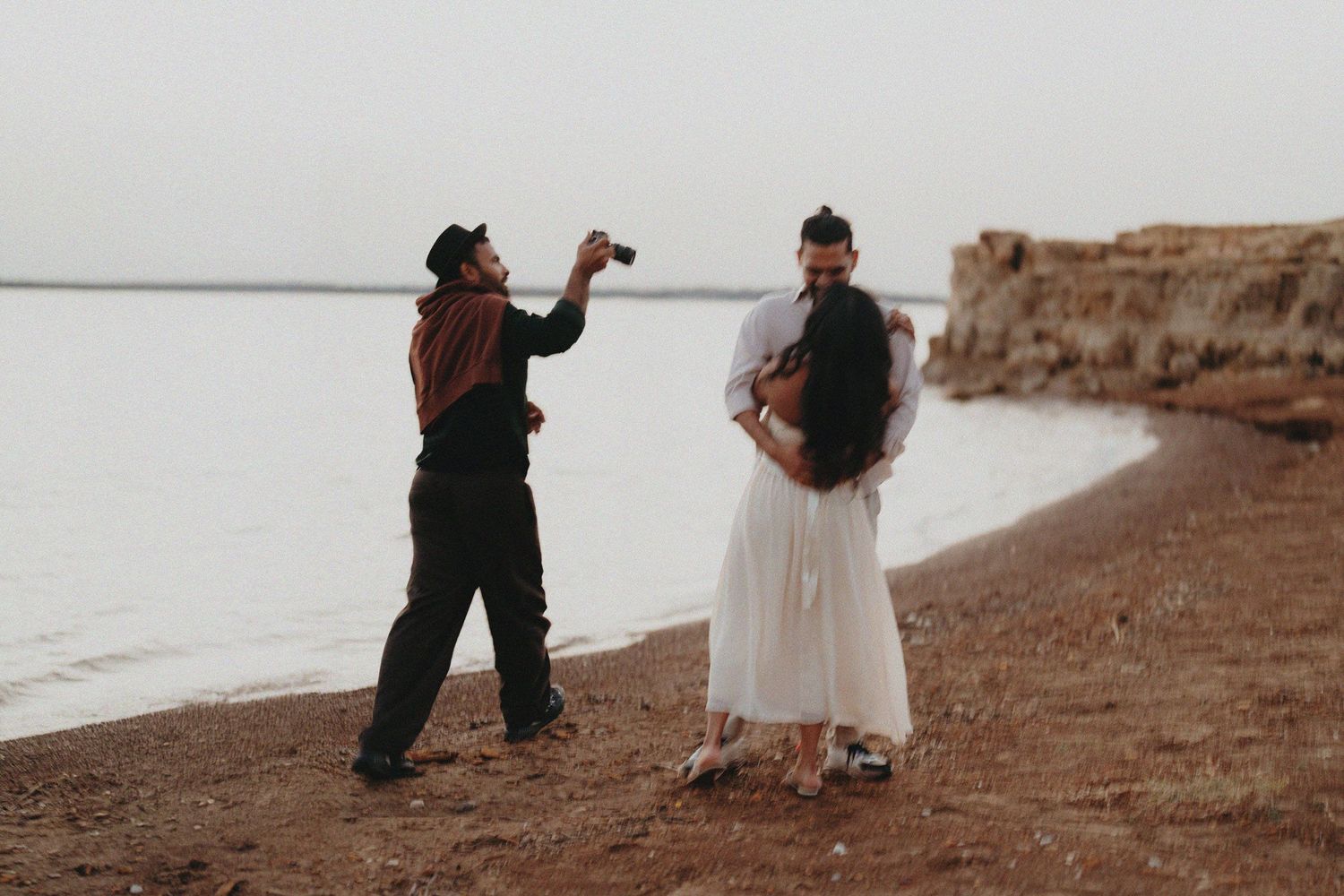 Photographer capturing a couple embracing on a rocky beach shoreline during an outdoor engagement or wedding photoshoot.