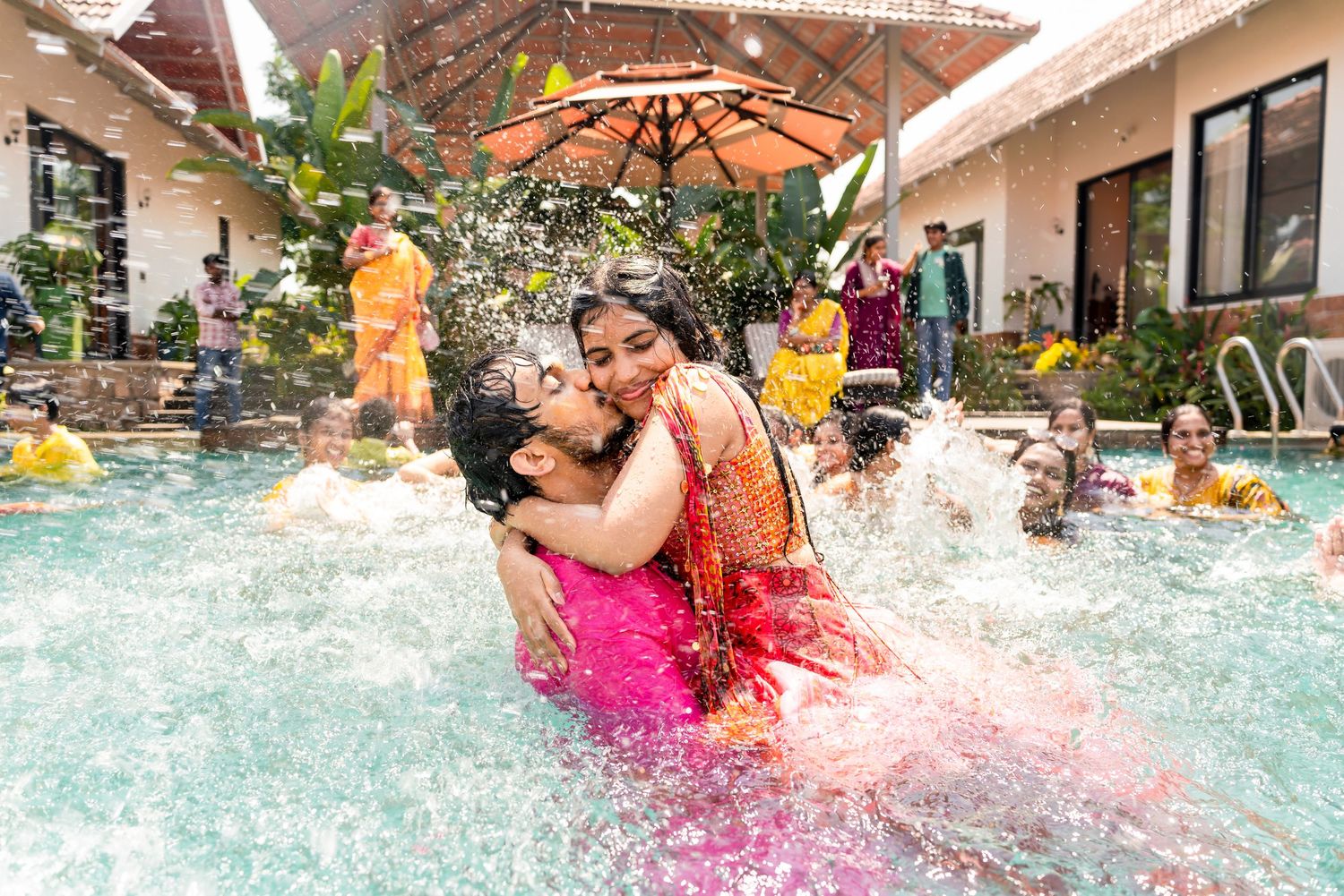 A bride and groom share a playful kiss while splashing in a pool during a vibrant Haldi ceremony.