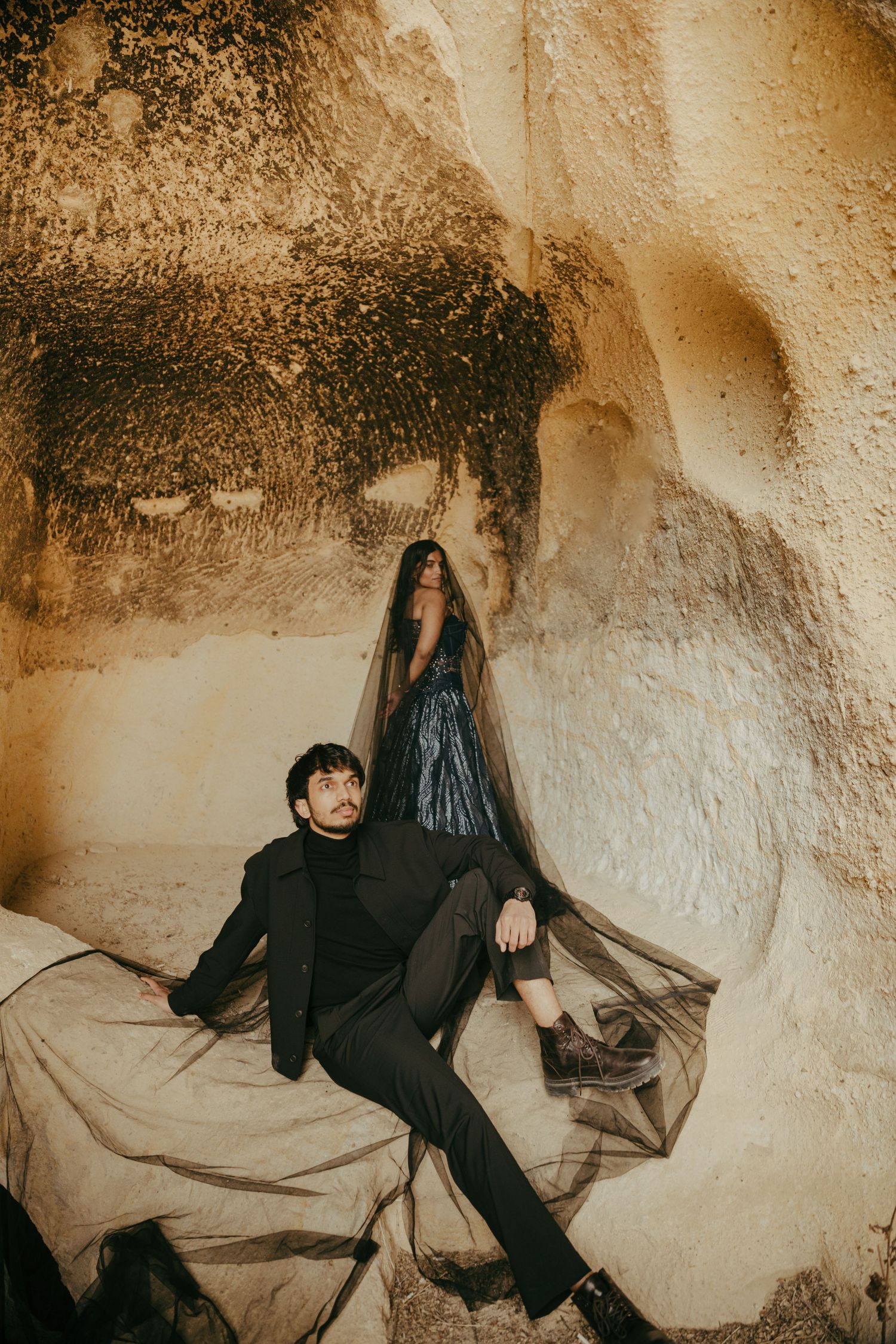 Couple in elegant black attire reclining against ancient cave rock formations with sheer tulle fabric.